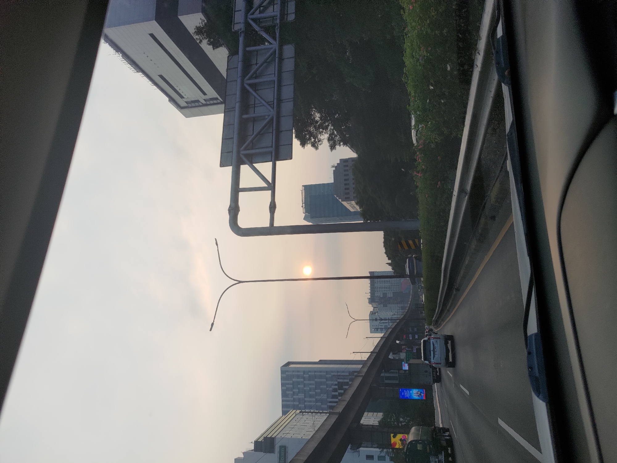The image depicts a view from a vehicle traveling on a multi-lane highway in South Jakarta, Indonesia, likely during late afternoon or early evening. The sky is overcast with a hazy, pale yellow hue, and a small, bright sun is visible near the horizon, suggesting either sunrise or sunset.
The highway features multiple lanes of traffic. In the foreground, the dashboard and windshield of the vehicle from which the photo is taken are visible. Ahead, several cars and trucks are driving on the highway. A white SUV is in the right lane, and a truck is in the left lane.
On the left side of the highway, an elevated train track structure runs parallel to the road, supported by concrete pillars. Adjacent to the train tracks, modern, multi-story buildings and some billboard advertisements are visible. One building on the far left has the text "team" on it.
On the right side of the highway, dense green foliage lines the road, with some pink flowers visible at the bottom. A large overhead road sign structure with metal supports and a sign panel is also present on the right. Beyond the foliage, more buildings, including a tall, modern skyscraper, can be seen in the distance. Streetlights are positioned along the highway. The overall impression is an urban highway scene within a bustling city.