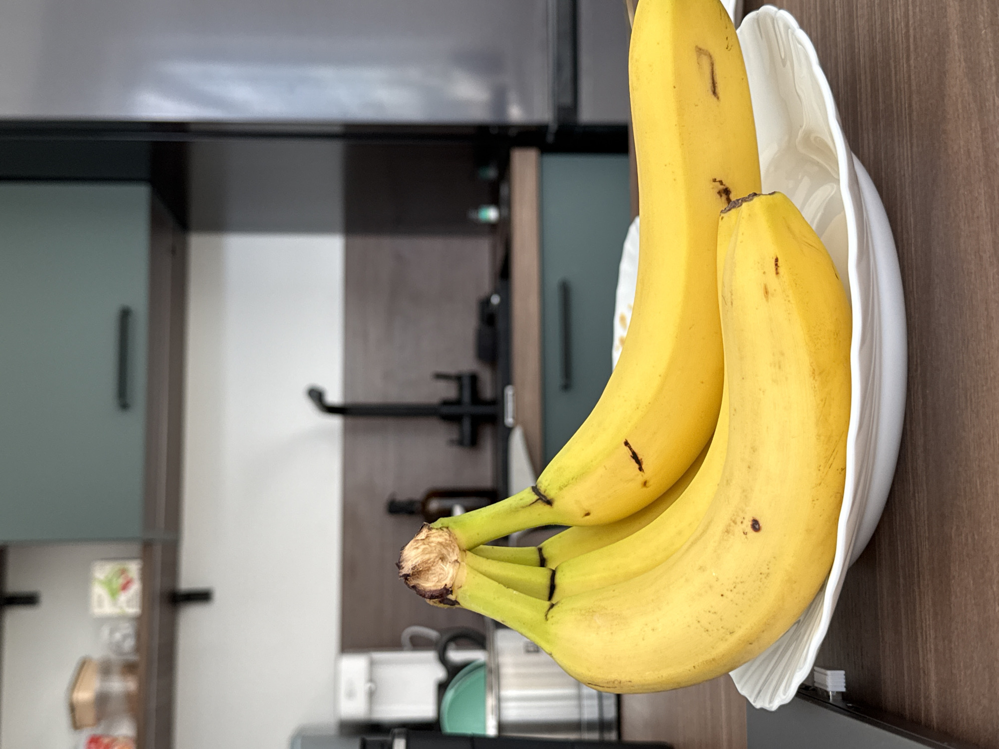 The image displays a close-up of a bunch of ripe yellow bananas resting in a white bowl. The bananas are the primary subjects, with varying degrees of ripeness indicated by small brown spots on their peels. The bowl has a scalloped edge and is positioned on a wooden surface.
In the background, the scene suggests a kitchen setting. Cabinets with teal-colored doors are visible, along with kitchen appliances like a faucet and what appears to be a refrigerator. The overall color palette is muted, with dominant tones of yellow from the bananas, white from the bowl, and browns and grays from the kitchen elements. The lighting appears to be artificial, typical of an indoor environment.
No people are visible in the image. The image seems to capture a moment of still life, focusing on the fruit and its immediate surroundings. The time of day is indeterminate. There is no visible text in the image. The location context of Saint Petersburg, Russia, is provided but not visually evident within the frame itself.