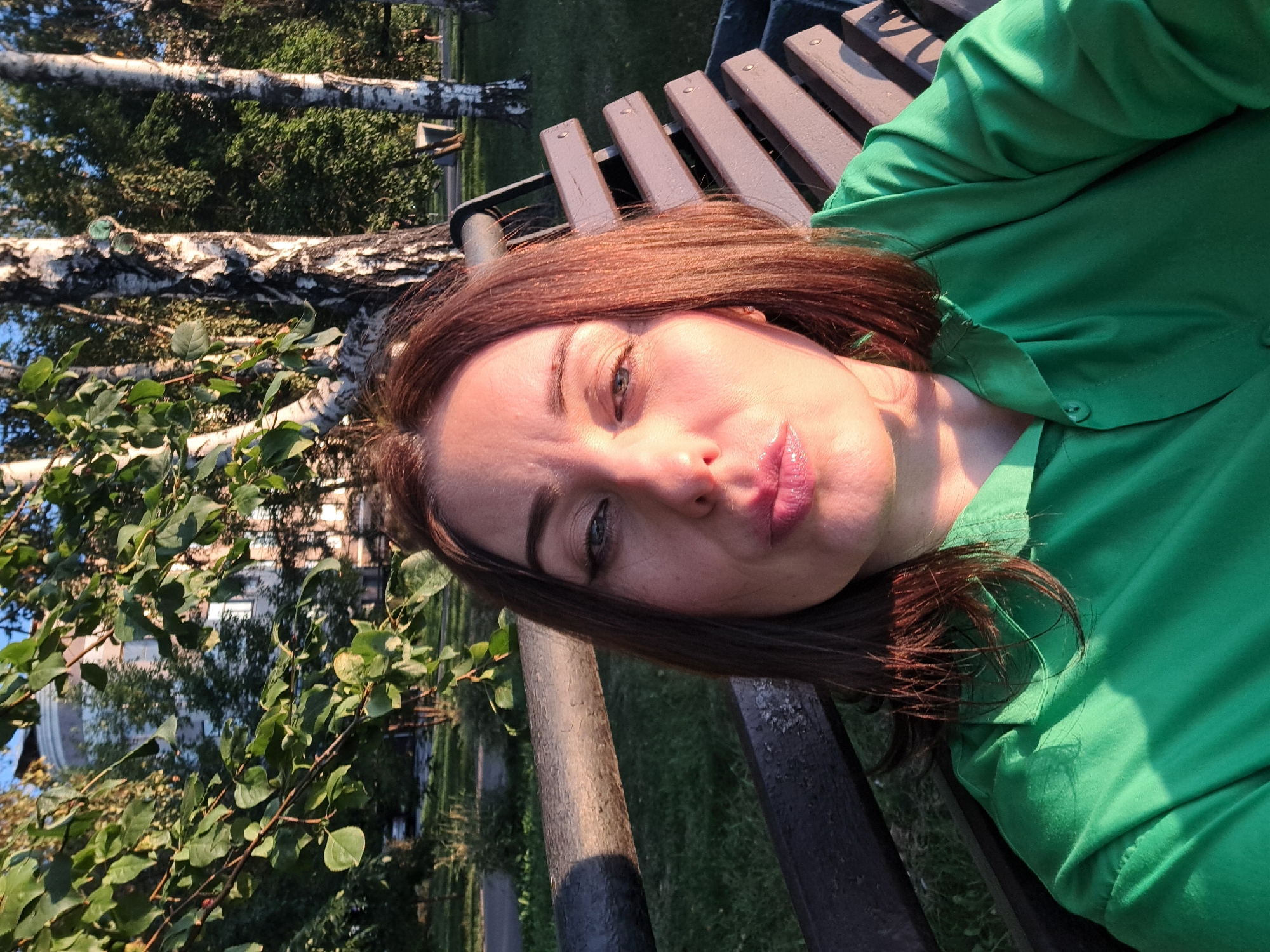 A medium shot selfie of a woman with auburn hair and makeup is taken outdoors in Irkutsk, Russia. The woman is wearing a green collared shirt. She is seated on a wooden park bench with a dark, possibly metal, armrest. The background includes trees, grass, and a building. The lighting suggests it is either early morning or late afternoon, as the sun is casting long shadows. The woman is looking towards the camera with her lips pursed.