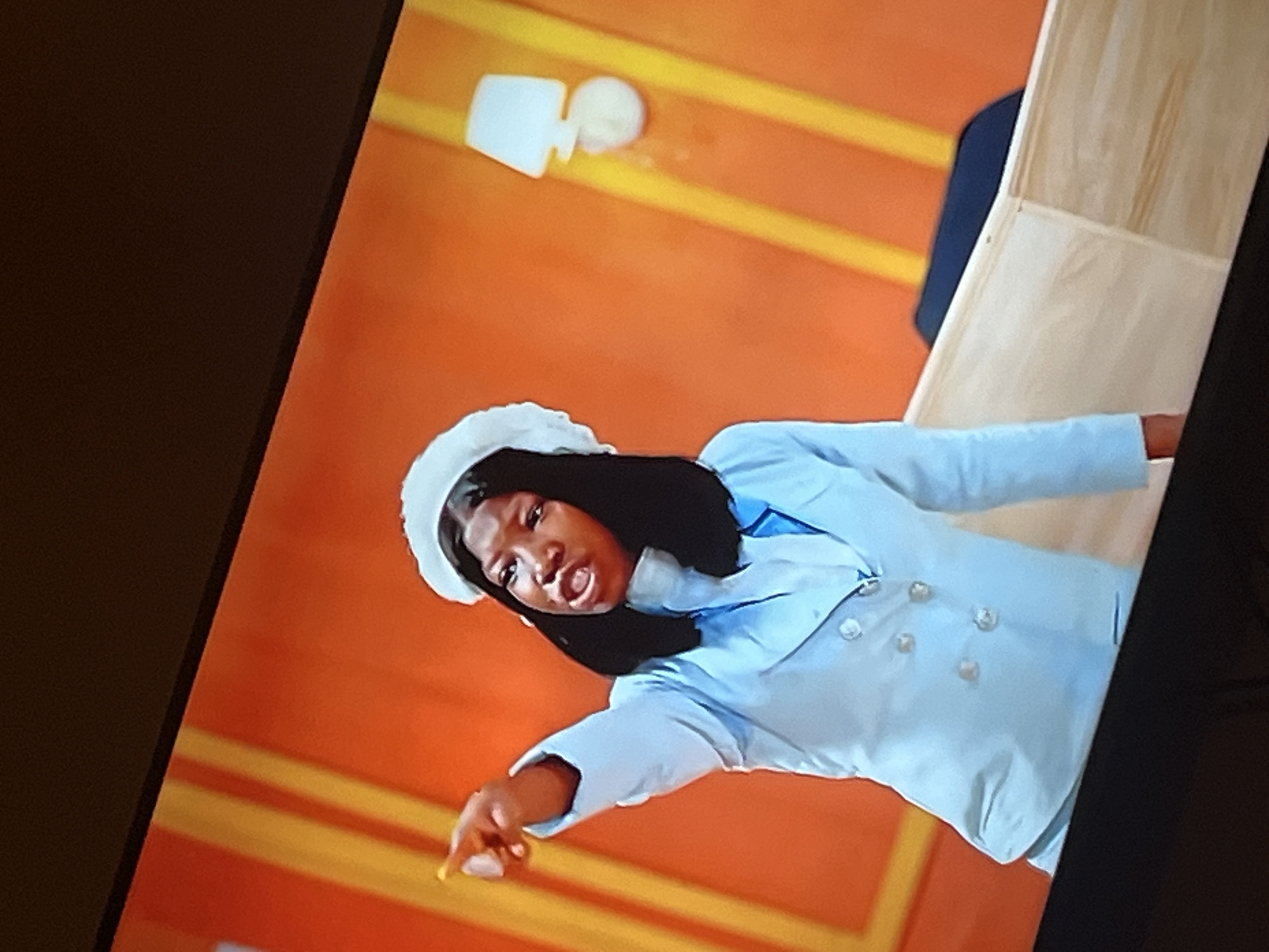 A young woman with dark hair and a white head covering is pointing her left index finger towards the left of the frame. She is wearing a light blue double-breasted jacket over a white turtleneck. The background is a vibrant orange wall with vertical yellow stripes. To the right of the woman, a white lamp is mounted on the wall. In the foreground, a light-colored wooden structure is partially visible. The image is a photograph taken from a slightly low angle, possibly of a television screen, given the dark border at the top and bottom of the frame. The scene is likely a studio set or a decorated interior space. The location in Gura Top, Nigeria is not discernable from the visual information provided.