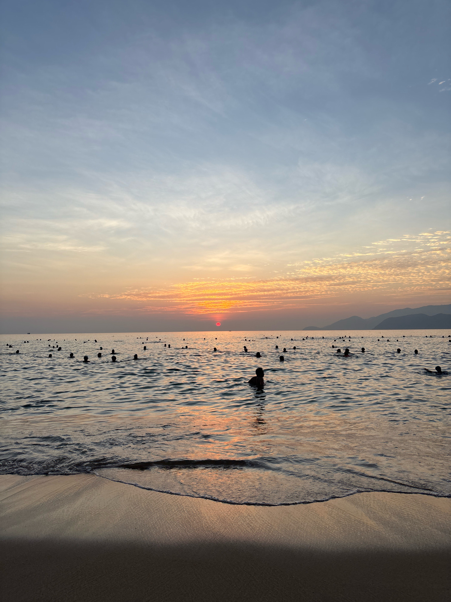 The image captures a sunset over the sea in Nha Trang, Vietnam. The sky is painted with hues of orange, pink, and blue, with scattered clouds. The sun, a vibrant red orb, is visible on the horizon, casting a reflection on the water. The sea is calm, with gentle waves lapping at the shore. Numerous people are swimming and playing in the water, their bodies silhouetted against the setting sun. The beach in the foreground is wet from the receding tide, with a gentle wave forming. In the distance, mountains are visible, their peaks softened by the twilight. The overall scene evokes a sense of peace and tranquility.