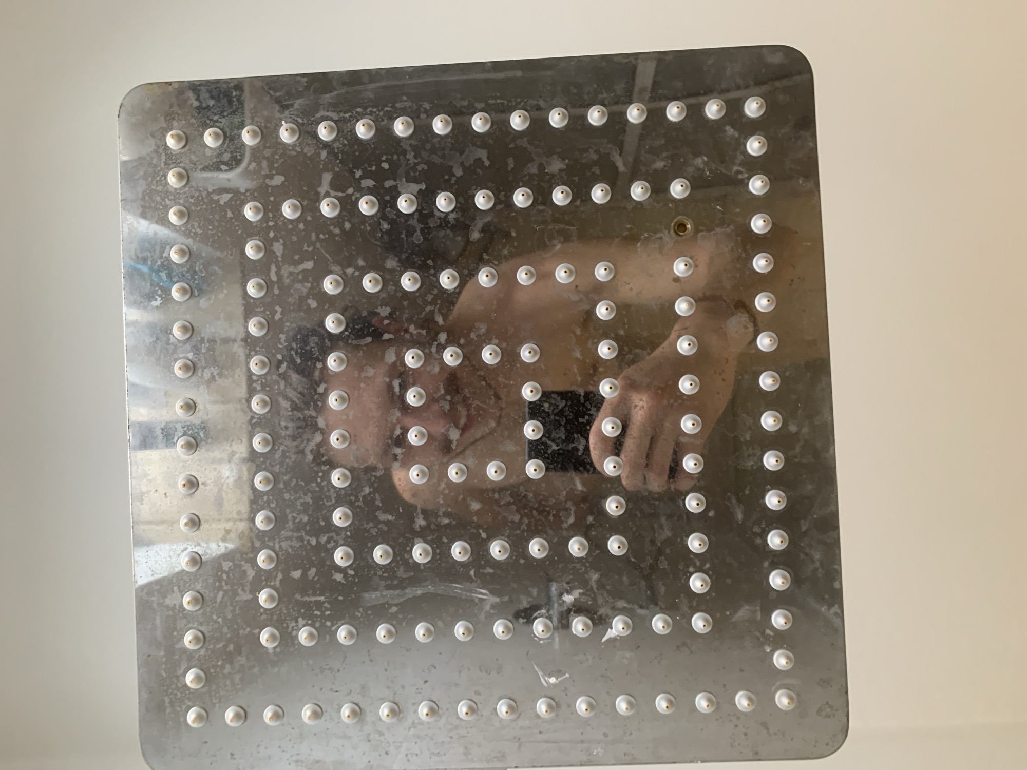 The image shows a square, metal showerhead with numerous small, circular nozzles. The showerhead appears to be dirty with water spots and soap residue. Reflected in the showerhead's surface is a person holding a smartphone, presumably taking the picture. The person is shirtless and their face is visible in the reflection. The background appears to be a plain, light-colored wall. There are no clues or information in the image that would allow identification of a location in the city of Tel-Aviv, Israel.