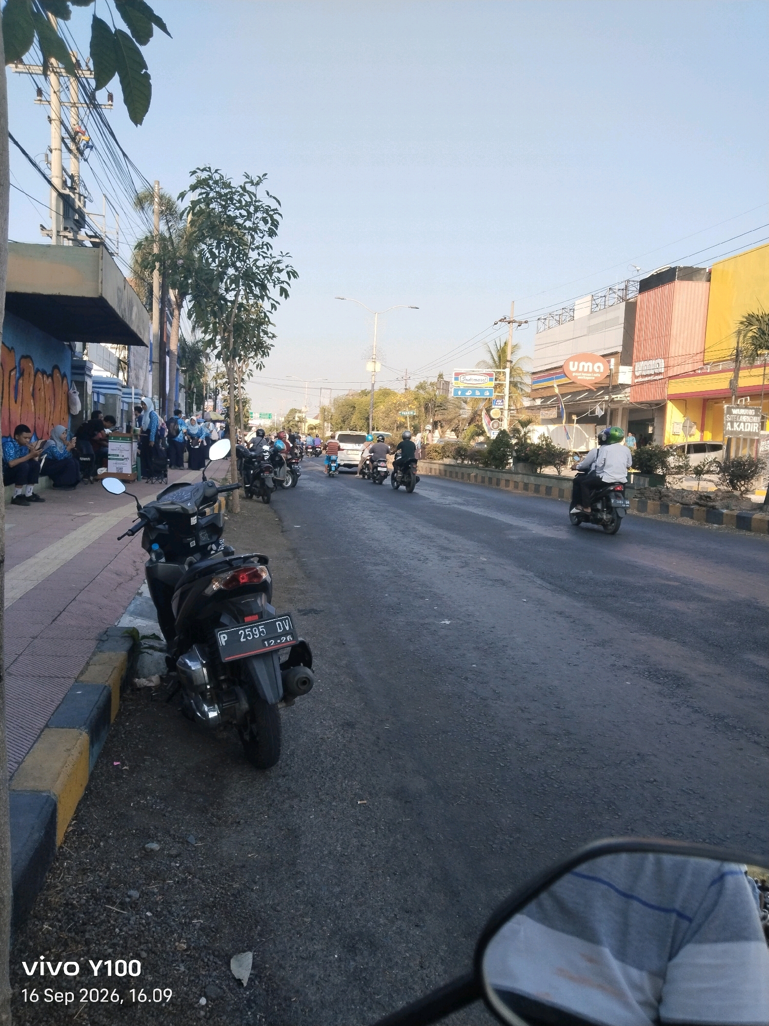 The image depicts a street scene in Situbondo, Indonesia, with several people and vehicles. On the left side of the road, a group of students in uniform are gathered, some sitting on a sidewalk and others standing. A black motorcycle is parked on the side of the road in the foreground, displaying a license plate that reads "P 2595 DV". The road is paved and appears to be a busy thoroughfare with other motorcycles and a car visible in the distance. Buildings line the right side of the street, including shops with colorful signage, one of which has "UMA" prominently displayed. The sky is clear and blue, suggesting it is daytime. Power lines and utility poles are visible along the street. The overall impression is a snapshot of daily life in a city.