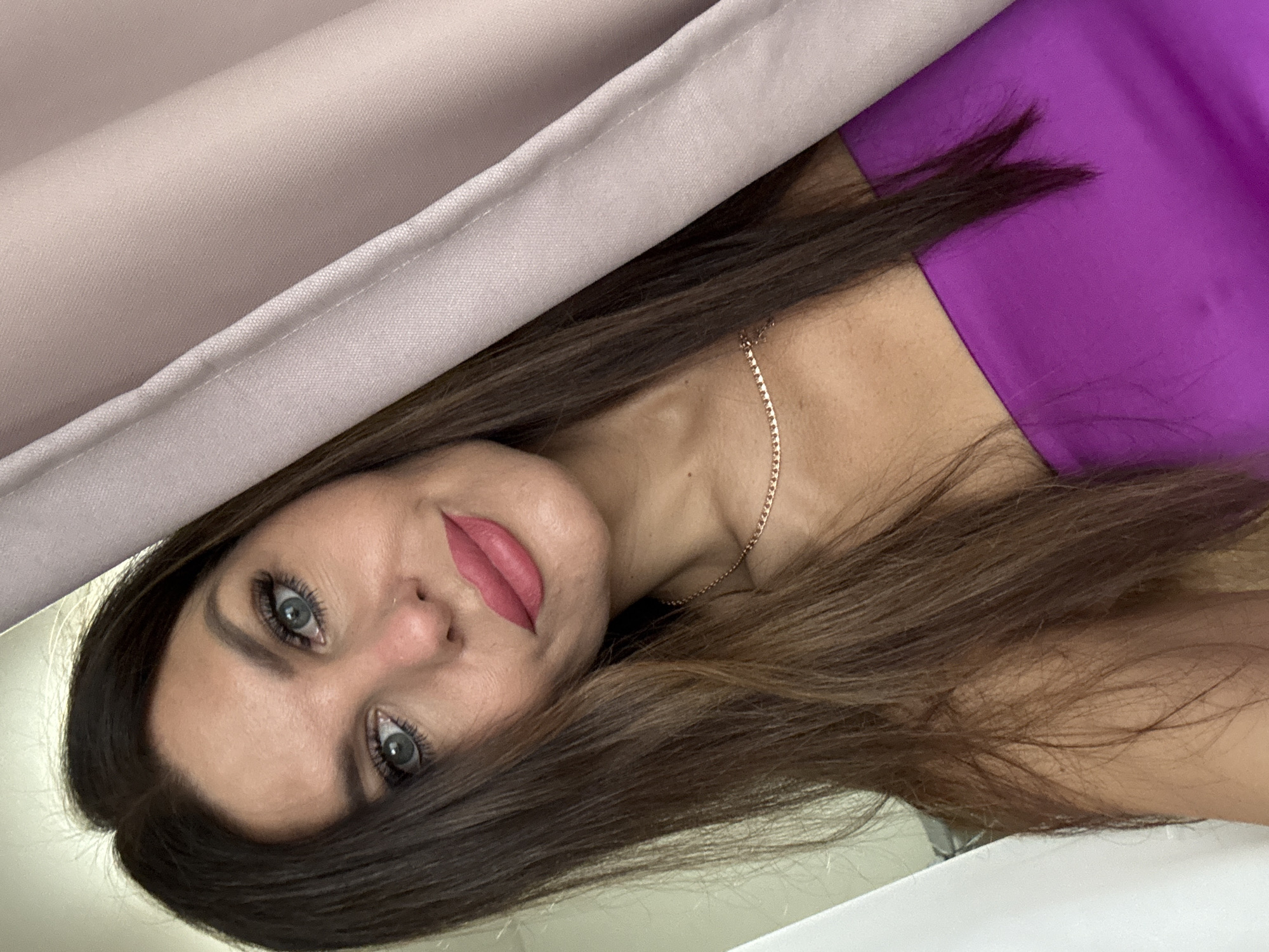 A medium shot captures a woman from the chest up, angled slightly to the right. She has long, dark brown hair framing her face, which is looking directly at the camera. Her eyes are blue-gray, with noticeable mascara and winged eyeliner. Her eyebrows are dark and well-defined. She is wearing a vibrant purple strapless top and a delicate gold chain necklace. Her lips are painted a matte pink shade. To the right of the woman, a light pink curtain hangs, partially obscuring her and the background. A white curtain or wall is visible on the left side of the frame. The image appears to be taken indoors, possibly in a home or dressing room, with soft lighting. No specific location in Chaikovsky, Russia is identifiable.