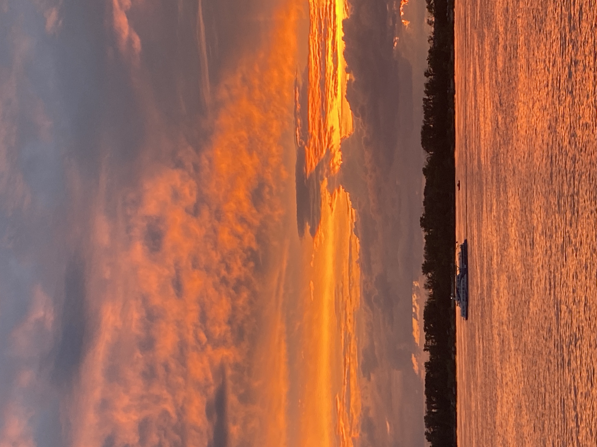 The image captures a vibrant sunset over a wide body of water, likely the Volga River, near Novokuybyshevsk, Russia. The sky is dominated by dramatic clouds, illuminated in intense shades of orange, red, and yellow, with darker grey clouds interspersed. On the water, a modern, multi-deck passenger boat is visible in the mid-distance, moving from left to right. A dark, tree-lined shore runs along the horizon. The water itself reflects the fiery colors of the sky, creating a shimmering, warm-toned surface. No people are clearly discernible on the boat or shore.