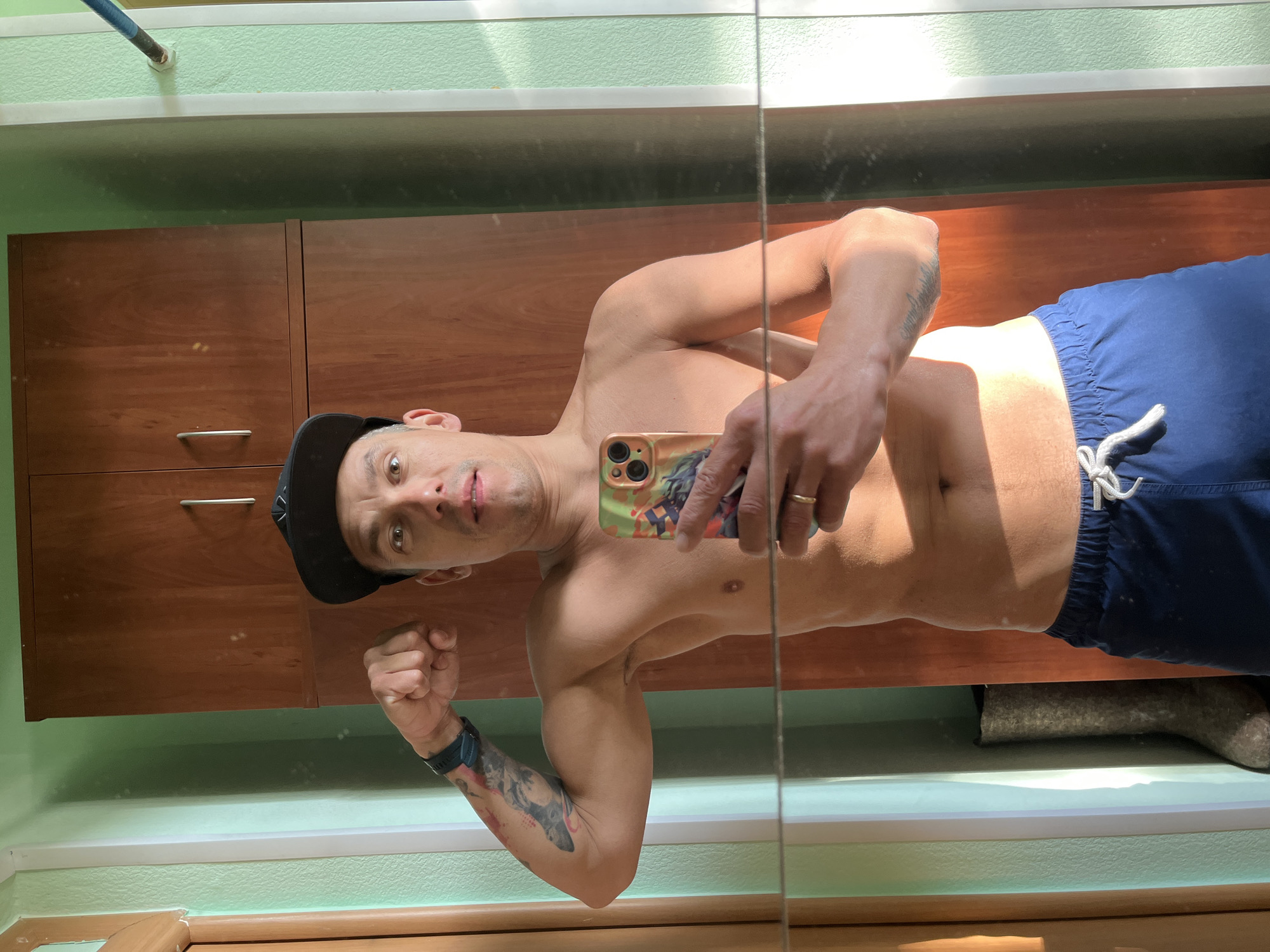 A shirtless man wearing blue shorts and a black baseball cap takes a selfie in a mirror. He is holding a smartphone in his right hand, and his left arm is flexed. A tattoo is visible on his left forearm. The background includes a wooden cabinet, green walls, and a window with sunlight streaming through. A felt boot is partially visible in the lower right corner. Based on the user's prompt, the location is assumed to be in the city of Chaikovsky, Russia.