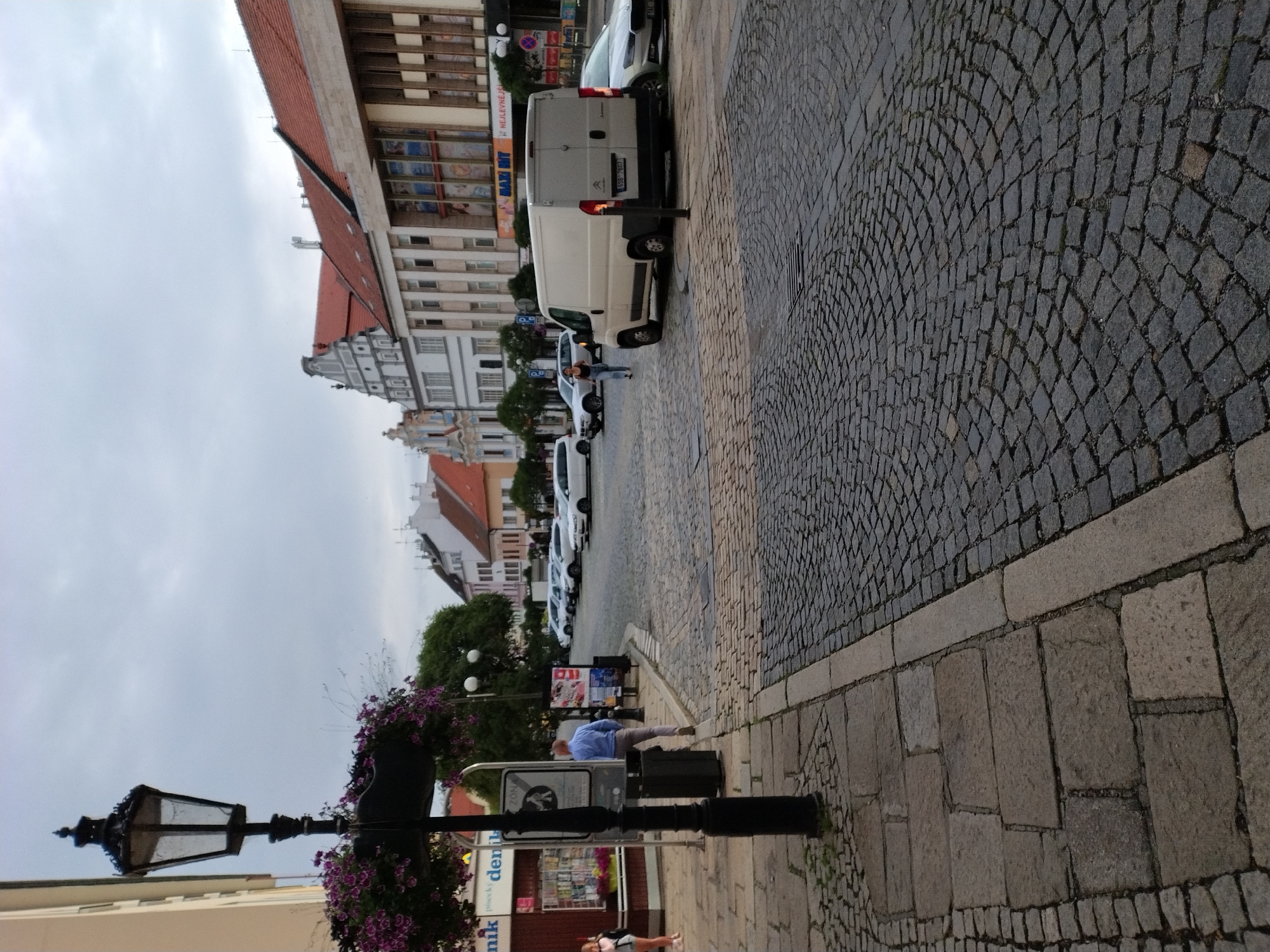 The image depicts a street scene in the city of Písek, Czechia, with a cobblestone road and sidewalk. In the foreground, a man wearing a light blue shirt and grey pants stands with his back to the camera, facing a trash can. To his left, a decorative lamppost with hanging plants is visible, along with a small kiosk. Further down the street, several white cars are parked, and a white van is in motion. The buildings lining the street exhibit traditional European architecture with red tile roofs. The sky is overcast, suggesting a cloudy day.