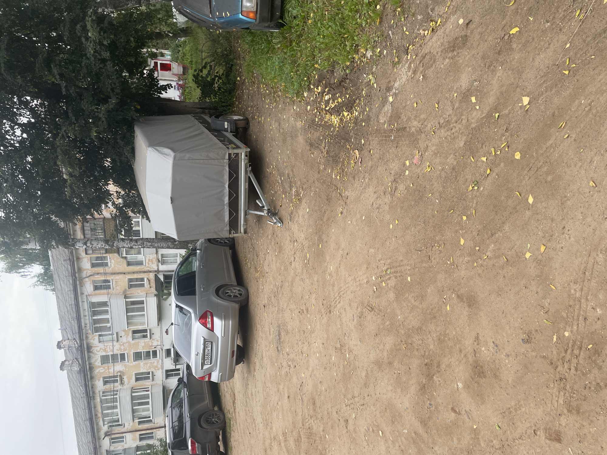 The image depicts a courtyard or parking area in front of a dilapidated apartment building in Chaikovsky, Russia. The building, likely constructed in the Soviet era, features peeling paint and numerous balconies. A silver sedan with a license plate reading "O368EH 159" is parked to the left, and a black SUV is parked behind it. A grey-covered trailer is positioned in the center of the frame, adjacent to a large tree. A portion of a blue car is visible on the right edge of the image. The ground is covered with dirt and scattered yellow leaves, suggesting an autumnal setting. The overall scene conveys a sense of urban decay and neglect.
