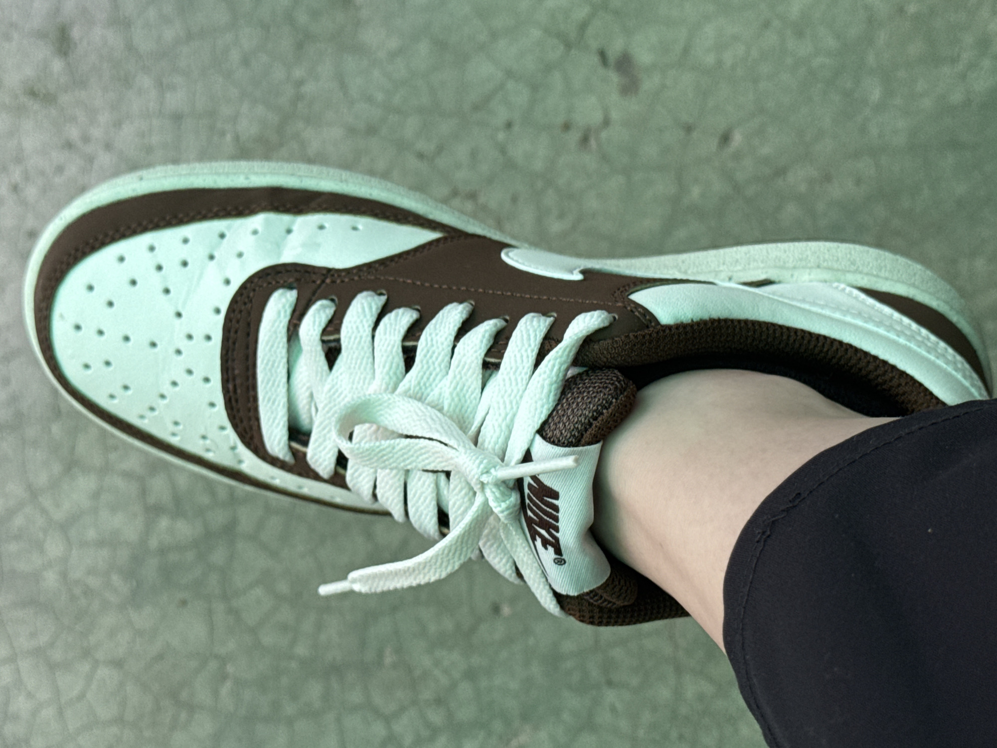 An overhead shot captures a person's left foot encased in a distinct Nike sneaker, resting on a light greenish-grey, textured surface, likely concrete or pavement. The shoe showcases a unique color combination of mint green and dark brown. Mint green panels form the toe box, which is perforated, and parts of the side and heel. Dark brown overlays accent the shoe, running along the lacing system, wrapping around the toe, and framing the heel.
The sneaker features white laces tied in a bow. The shoe's tongue, visible at the ankle, is white with the "NIKE" logo and swoosh printed in dark brown. A portion of the wearer's bare ankle is visible, along with the cuff of a dark, possibly black, garment like pants or leggings, which has minor fraying along its edge.
The lighting is bright and even, suggesting it is daytime, potentially outdoors. There are no harsh shadows, indicating an overcast day or indirect light. No specific activity is taking place, and no emotions are discernible from this close-up view. The background is slightly blurred, keeping the focus on the shoe. While the location context of Govedare, Bulgaria is provided, the image itself does not contain specific geographical cues.