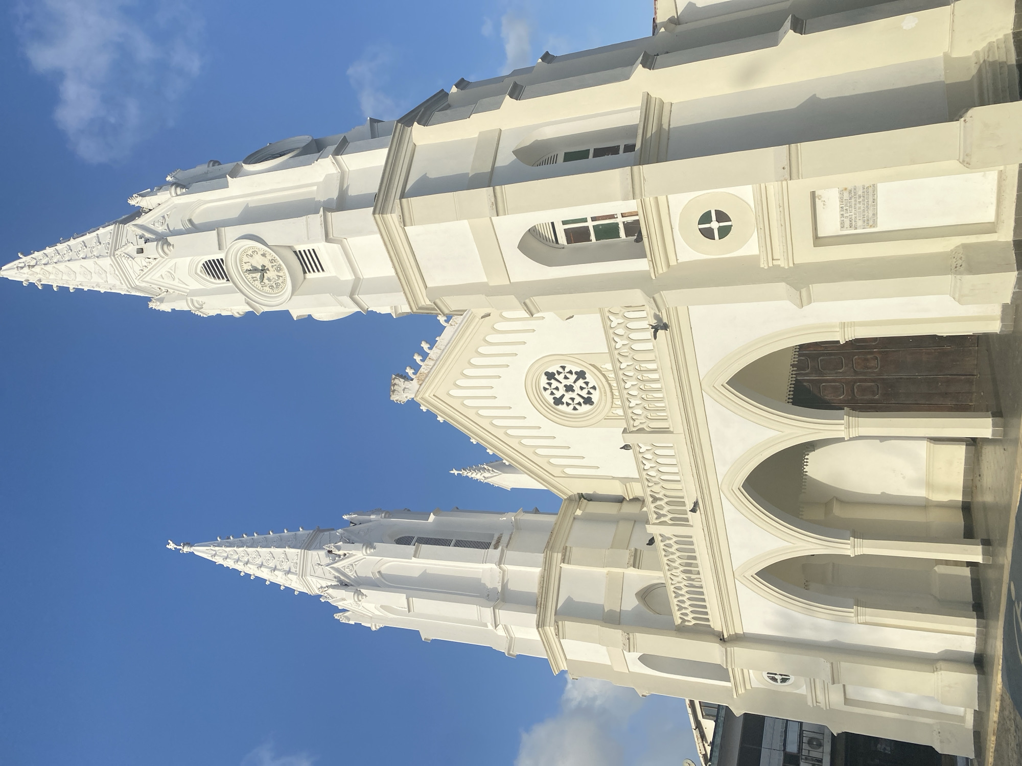The image shows the Iglesia de San Juan Evangelista, a historic white-painted church located in Juan Griego, Venezuela. The church features two prominent, intricately designed steeples that reach into a clear, blue sky with scattered clouds. The building exhibits a neo-Gothic architectural style with pointed arches, decorative windows, and ornate detailing. The main entrance is framed by a series of arched columns and includes a large, dark wooden door. A plaque is visible on the building's facade. No people are visible in the frame.