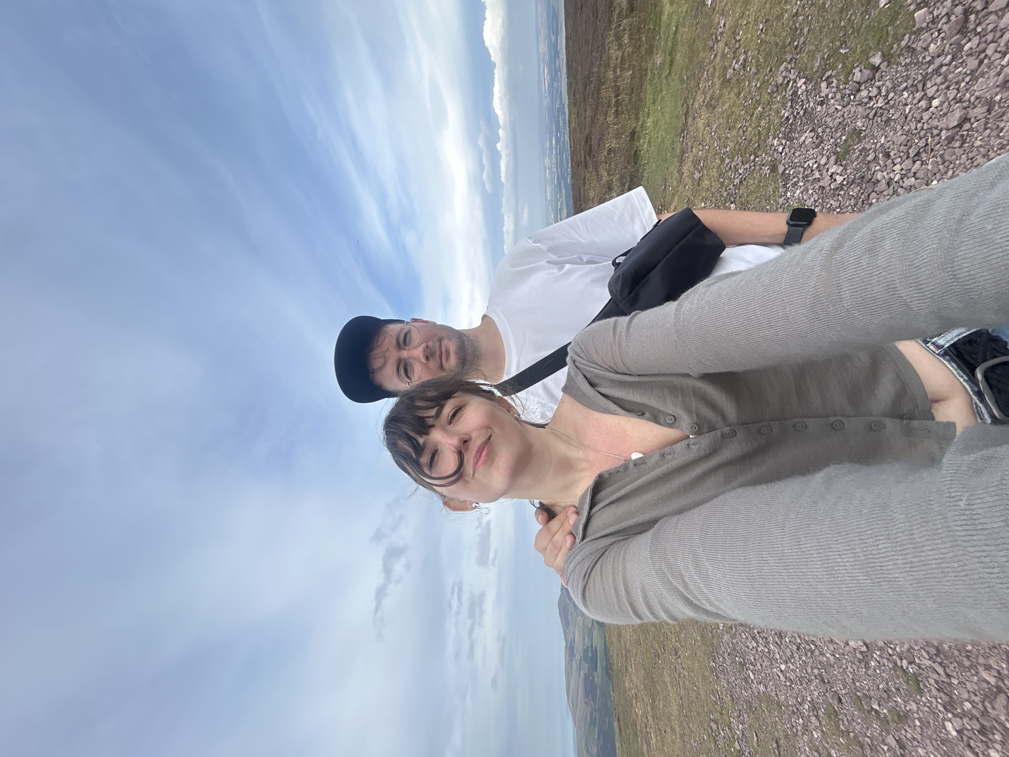 Two individuals, a woman in the foreground and a man behind her, are taking a selfie on a rocky, grassy hillside with a cloudy sky. The woman has dark hair with bangs and is wearing a light brown, button-up cardigan and jeans. She is smiling and looking at the camera, with her arm around the man's shoulder. The man is wearing a white t-shirt, a black baseball cap, and glasses. He has a black bag slung across his body. The background shows rolling hills and a distant view of what appears to be a town or city. The location is identified as being in the vicinity of Penicuik, United Kingdom.