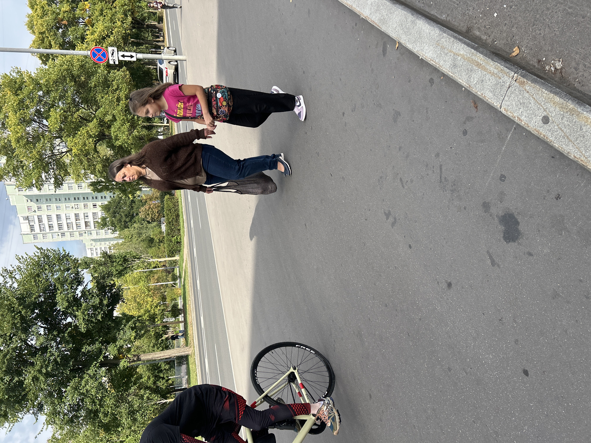 The image depicts a street scene in Moscow, Russia. In the foreground, a person is riding a bicycle from left to right. The cyclist is wearing black leggings with a red pattern and a black top. The bicycle has a light-colored frame and black wheels.
To the right of the cyclist, a woman and a young girl are walking from left to right, towards the viewer. The woman is wearing a brown cardigan over a lighter-colored shirt and dark blue jeans. She is carrying a dark mesh bag in her left hand. The girl is wearing a pink t-shirt with a graphic design and black pants. She has a colorful fanny pack around her waist.
In the background, trees with green and yellow foliage line the street. Several buildings are visible, including a tall, light-colored residential building with many windows. A street sign with a red circle and a white arrow pointing down is visible on a pole. A silver car is parked on the right side of the street. The pavement is asphalt, with a concrete curb on the right side of the image. The sky is overcast.