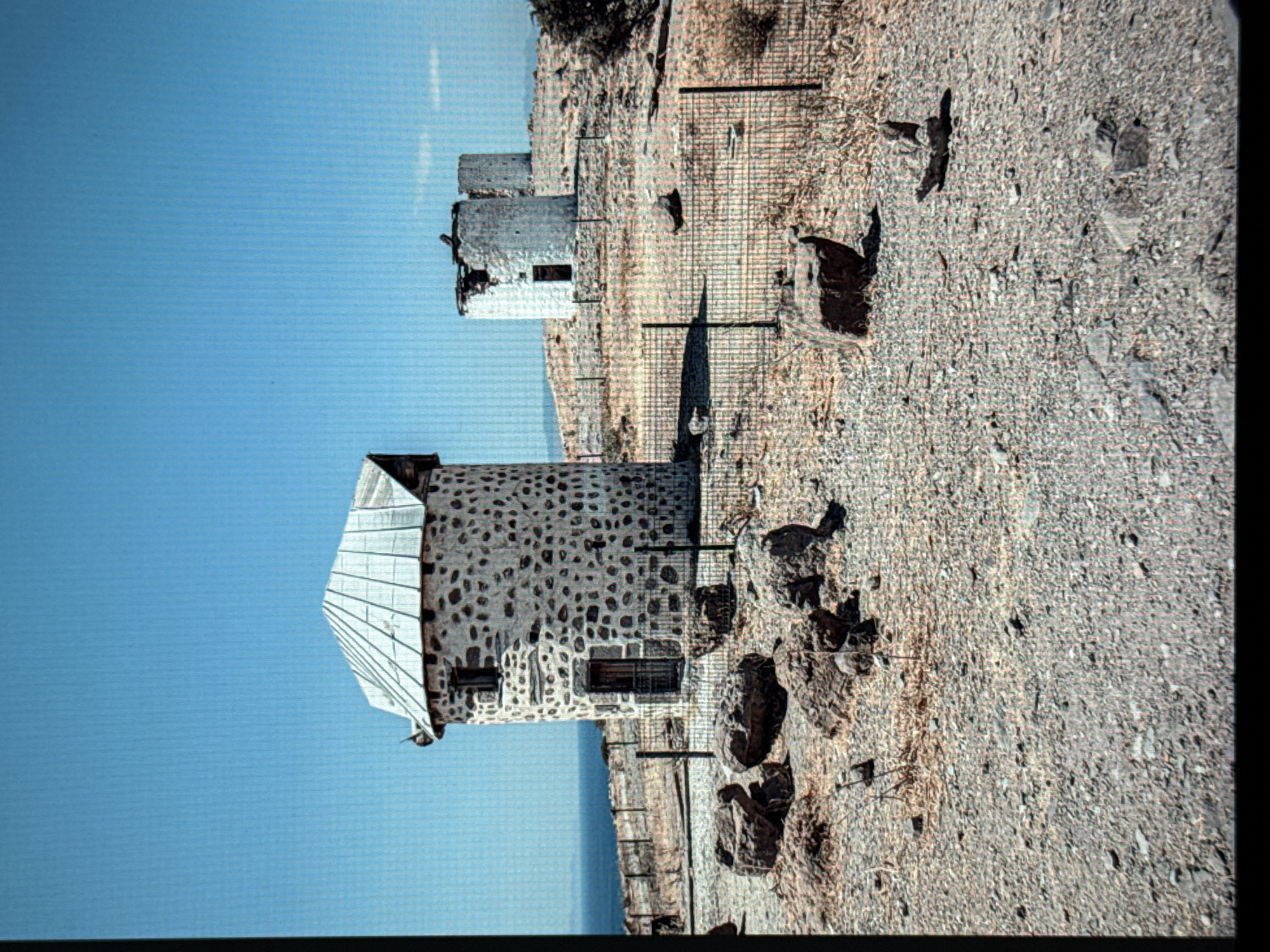An outdoor medium shot captures a stone-built windmill on a rocky, arid hillside under a clear blue sky. The windmill, made of irregularly shaped grey stones, has a conical roof with metal sheeting and a small, square window. A dark wooden door is set into its facade. In the background, two more similar, though more dilapidated, windmills are visible on the ridge. A black metal fence with vertical posts separates the foreground from the windmills. Large, dark rocks are scattered on the dry, gravelly ground in the foreground, with sparse, dry vegetation. The sea is visible in the distance on the left, meeting the horizon under the bright sky. The overall impression is one of historical ruins in a remote, sun-baked landscape.