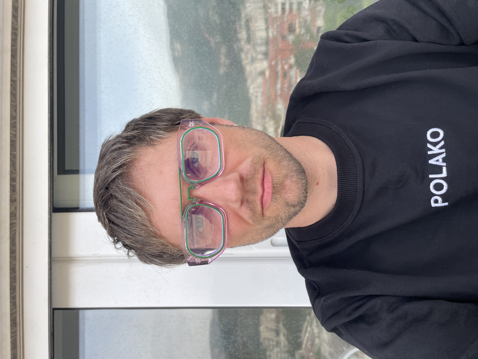 The image shows a man wearing glasses and a black sweatshirt with the word "POLAKO" written in white letters on the right side. The glasses have a combination of pink and green frames. The man has short, graying hair and a light complexion. The background appears to be a window with a view of a city and mountains. The city appears to be Budva, Montenegro.