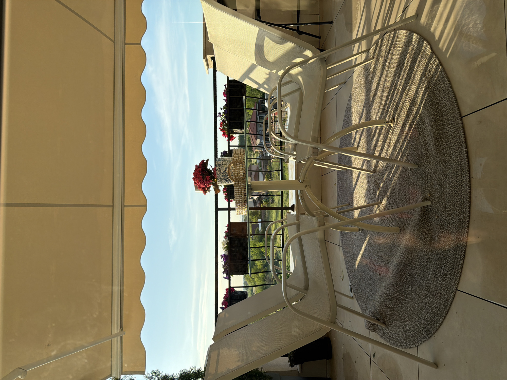 The image shows a patio scene in Plovdiv, Bulgaria. The patio is covered by a retractable awning. In the center, there is a round glass-topped table with a bouquet of red flowers and a decorative basket on top. Four white metal chairs are arranged around the table, two are positioned at the left edge of the frame and two others at the right. Beneath the table and chairs is a round, gray woven rug. The patio overlooks a green landscape with houses and trees. Balcony flower boxes with red and purple flowers line the railing. The sky is clear and blue.