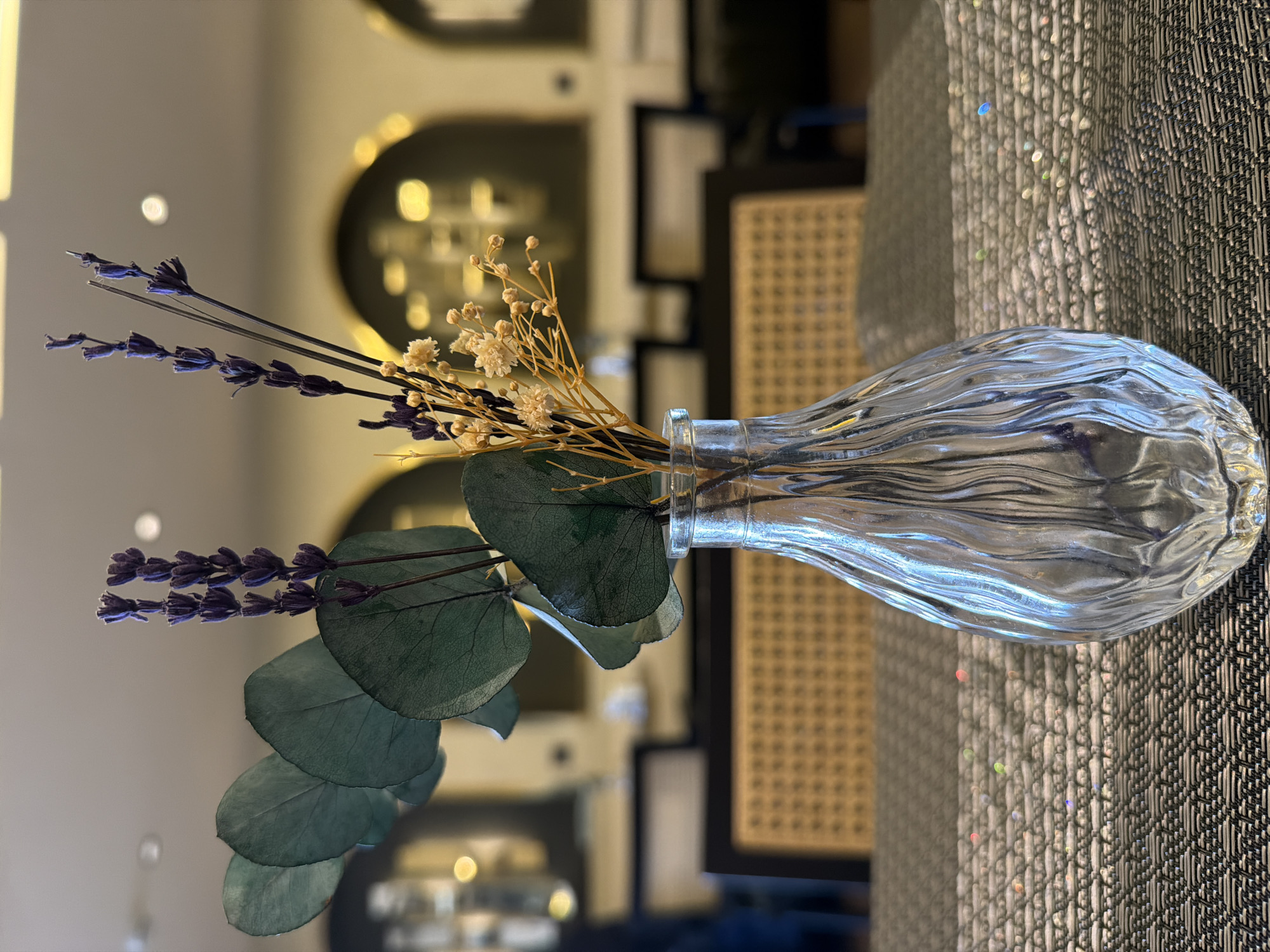 A slightly blurred, medium shot depicts a clear glass vase containing dried flowers and eucalyptus branches, laying on its side, likely on a table. The vase is filled with violet lavender flowers, small white dried flowers, and round, green eucalyptus leaves. The table has a textured, gray woven surface that reflects light. Behind the vase and flowers are blurred interior elements, including a woven-back chair and decorative archways with gold accents on a muted backdrop. The location is unspecified.