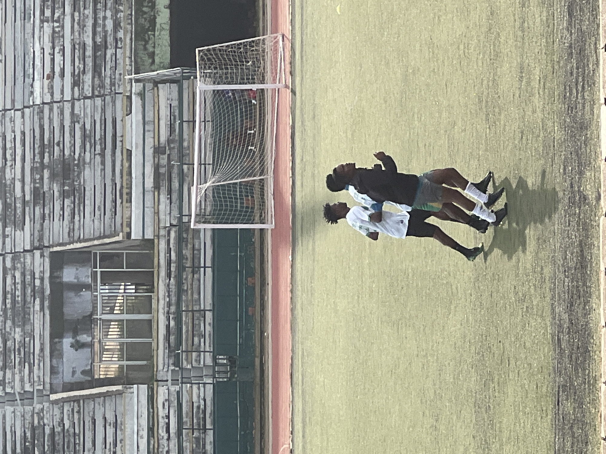 In the city of New Stadium, Nigeria, three young men are captured in mid-stride during what appears to be a football training session on a grassy field. They are running from left to right, their bodies in motion, suggesting active training. The background features a weathered stadium with tiered seating, a soccer goal with a net, and a running track. The lighting indicates an outdoor setting, likely during daylight hours. The scene is devoid of any specific interactions between the individuals, each focused on their individual movement across the field.