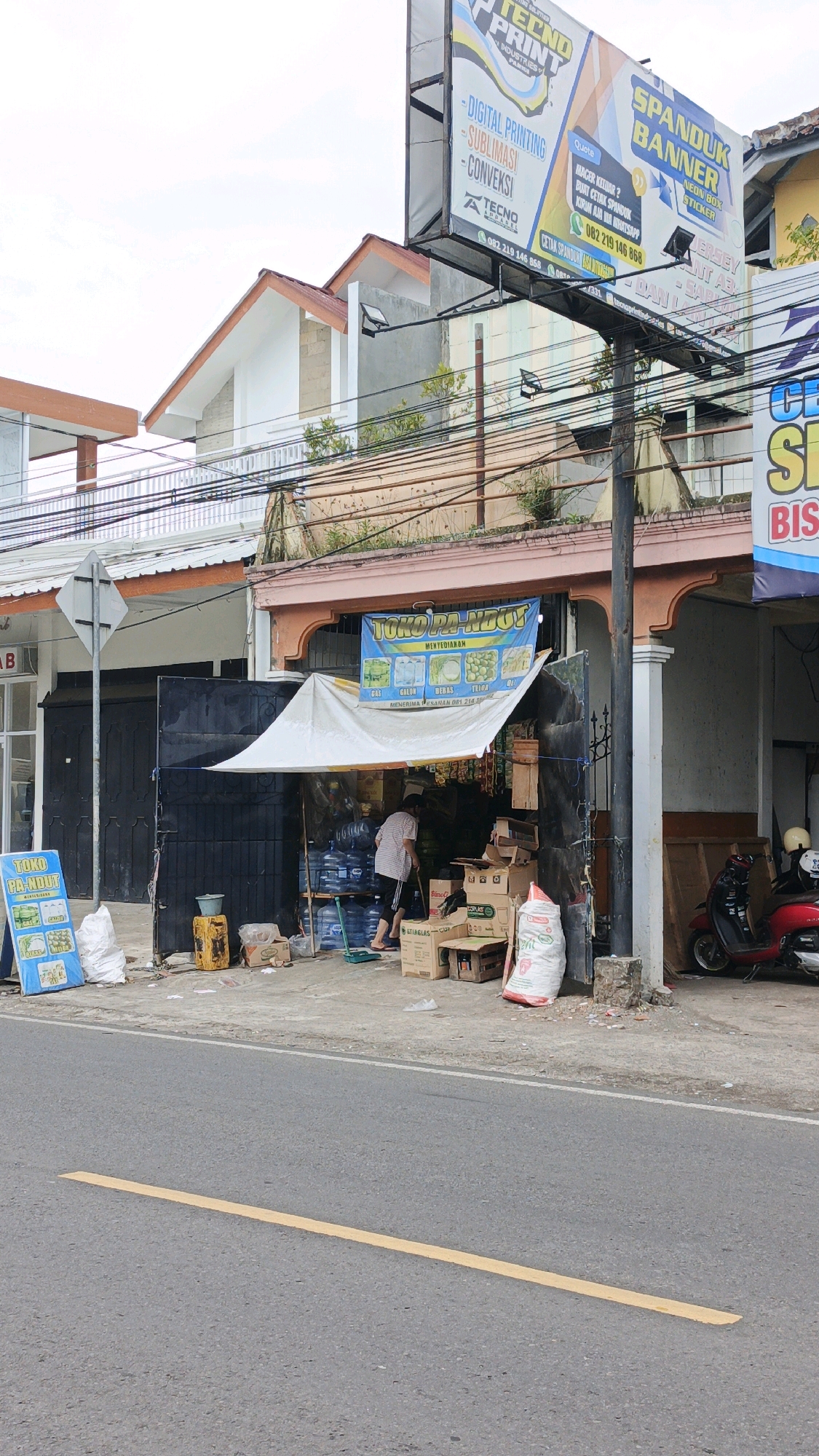 The image depicts a street scene in Desa Karangbenda, Indonesia, featuring a small shop and its surrounding area. In the foreground, a paved road with a yellow line is visible. To the left, a sign for "Toko Pa' Ndut" displays images of gas, water, ice, and snacks. A person in a white and black outfit is seen tending to bottled water and boxes inside the shop, which is covered by a white awning. Various items, including large plastic water containers, cardboard boxes, and bags of what appear to be goods, are arranged outside the shop. A yellow jerrycan is also present. In the background, buildings with tiled roofs and electrical wires overhead are visible, along with a large billboard advertising "TECKNO PRINT INDUSTRIES" and "SPANDUK BANNER". A red scooter is parked to the right of the shop. The sky is overcast.