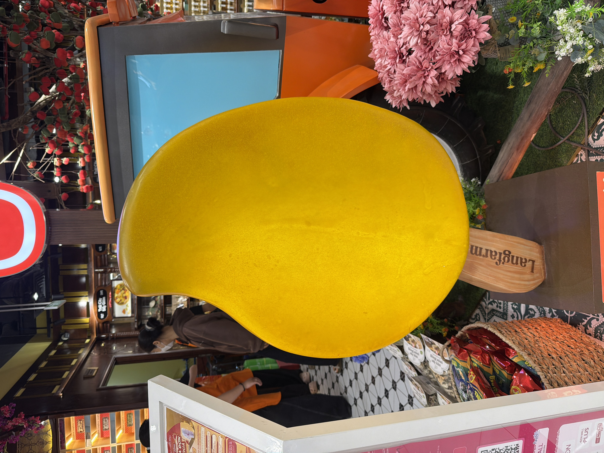 The image depicts a decorative display, likely in a commercial setting, featuring a large, yellow, mango-shaped object as the central focus. This object is mounted on a wooden stand that reads "Langfarm". Behind the mango, an orange and grey vehicle-like structure with a blue screen is visible. To the right of the mango, a cluster of pink flowers adds to the scene's aesthetics. In the background, shelves with products, hanging red decorations resembling fruit, and a tiled floor suggest an indoor retail or exhibition space. A few individuals are partially visible in the background, one wearing a brown hoodie and another in an orange garment. The overall impression is a vibrant and visually engaging display, possibly promoting a brand or product, and it is highly likely situated in a shopping mall or a similar establishment within Da Nang, Vietnam, given the presence of "Langfarm" which is a known Vietnamese brand.