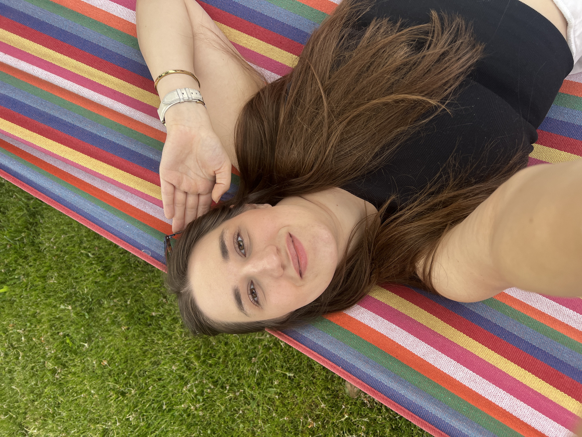 A woman lies in a multicolored striped hammock. She is wearing a black top and has long brown hair. She has a watch and bracelet on her left wrist. The hammock is suspended over green grass. The location is possibly Busmantsi, Bulgaria.