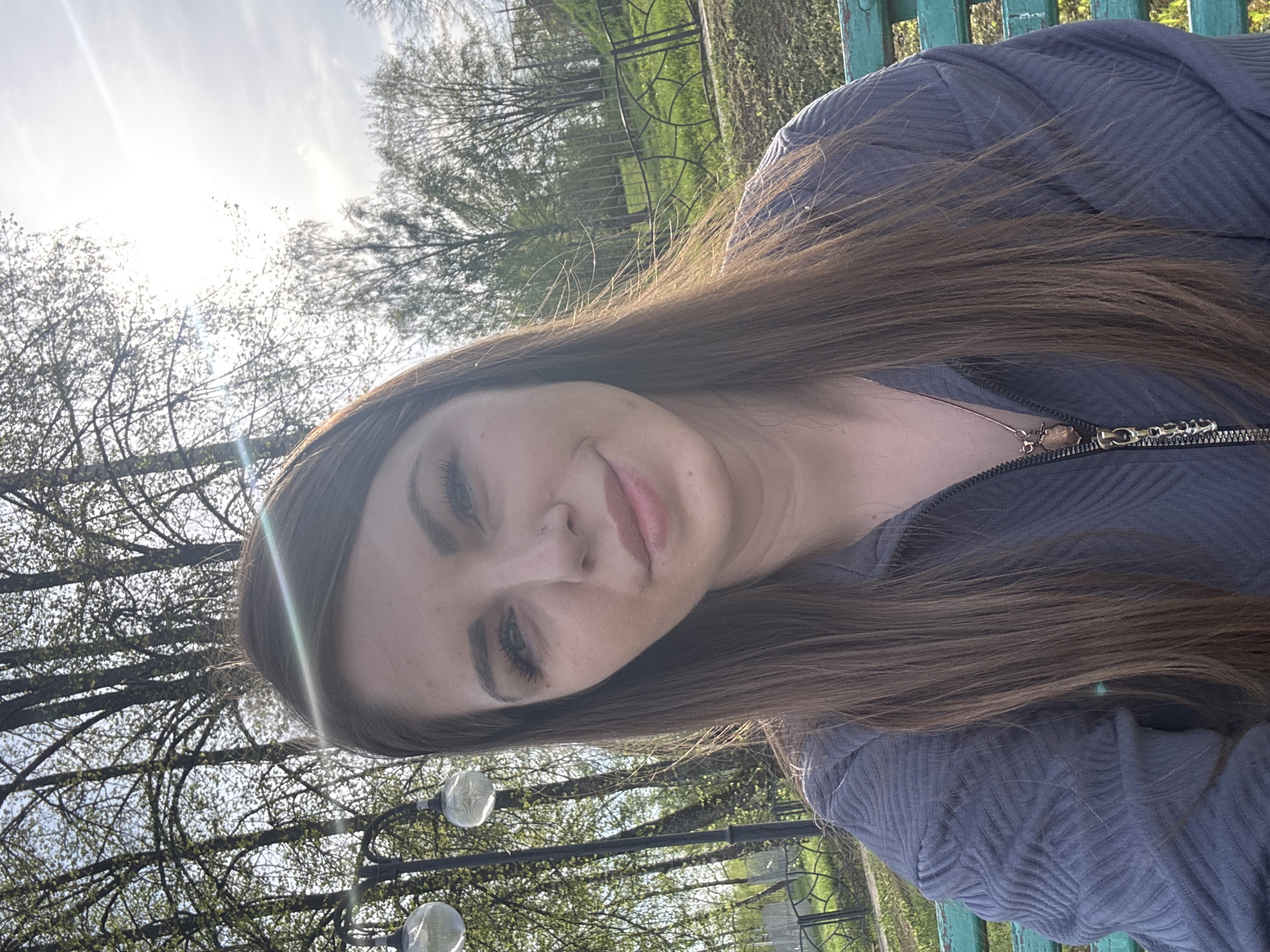 A young woman with long brown hair is sitting on a green park bench outdoors. She is wearing a zipped-up, gray-blue jacket with a textured pattern. A necklace with a pendant is visible. The background features trees, some with new spring leaves, and a wrought-iron fence. Street lamps are also present. The location is likely Shevlyakovo, Russia.