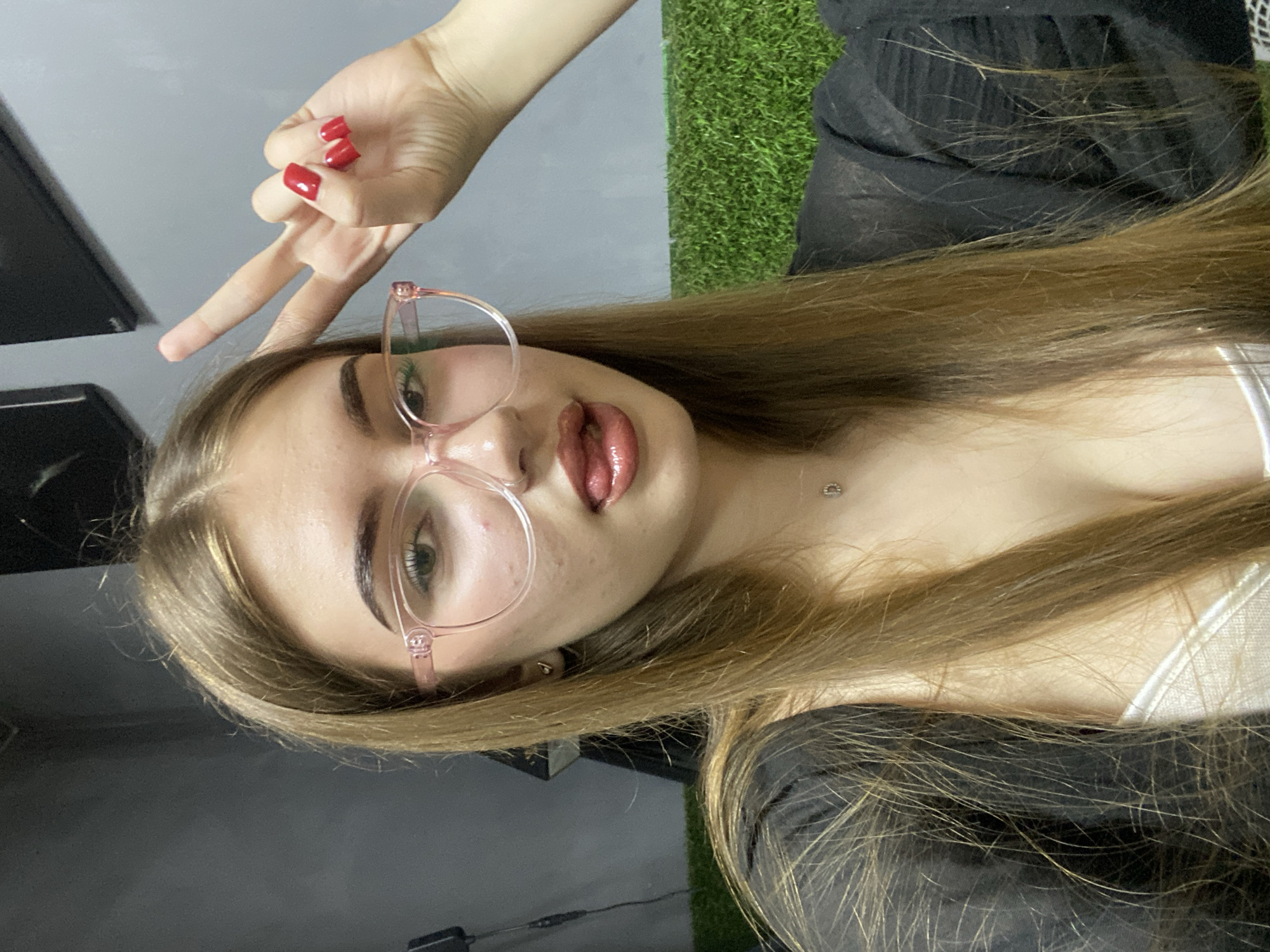A young woman with long, light brown hair and glasses is shown in a selfie. She is wearing a black sheer top over a white camisole, and a small pendant necklace. She is sticking her tongue out slightly and making a peace sign with her right hand. The background appears to be an indoor area with a grey wall and what looks like artificial grass on the lower right. There are no visible elements that would place this image specifically in the city of Дьяково, Russia.