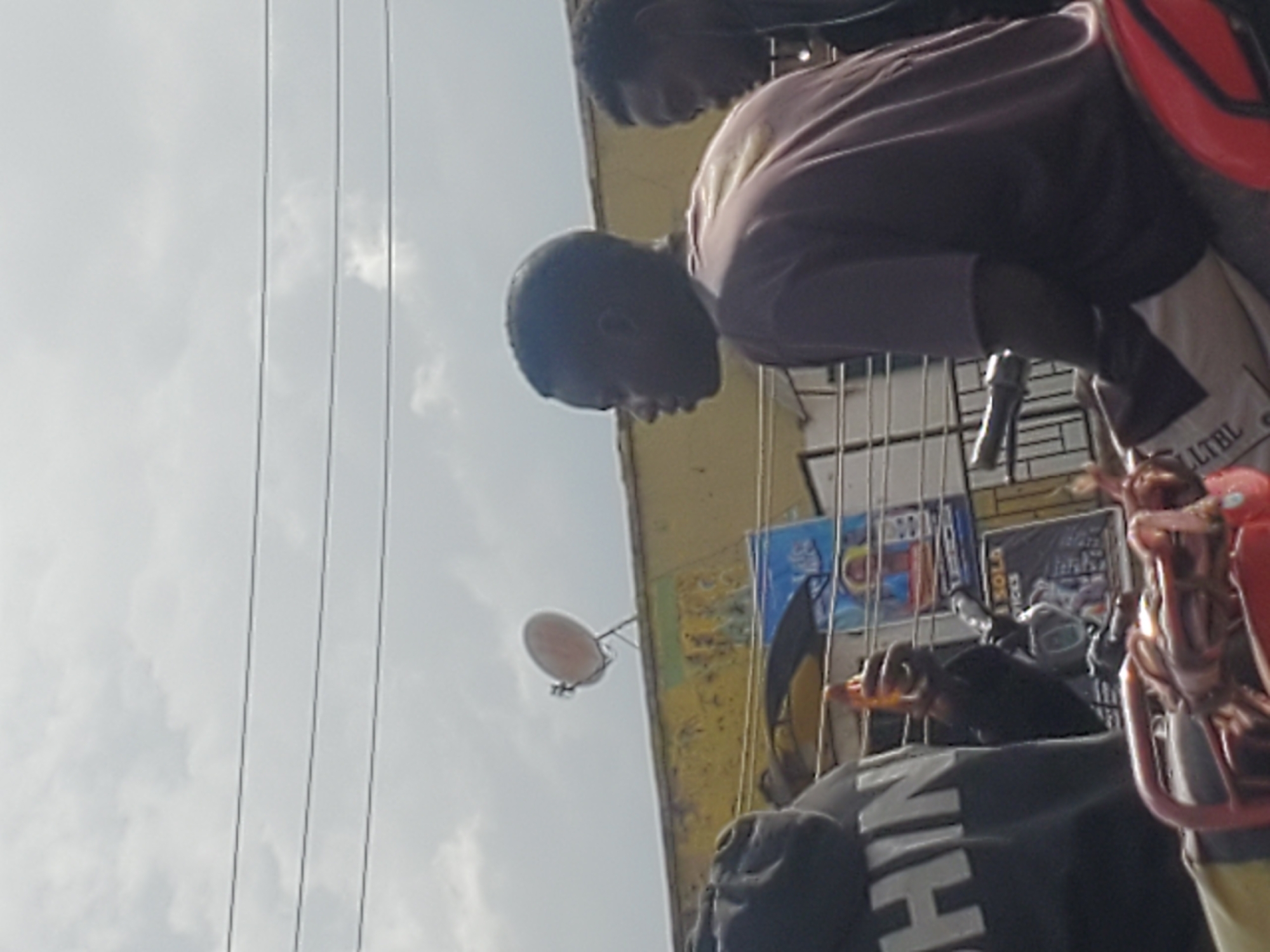 In Gwong, Nigeria, a cloudy sky looms above a street scene. Three individuals are visible, two of whom appear to be riding a motorcycle. The person in the foreground on the left wears a black hoodie with white lettering that partially reads "SHIN". This individual is holding an orange object in their right hand. To the right, a young man in a dark purple t-shirt is seated, looking to the left. Behind him, another individual, partially obscured, wears headphones and looks towards the right. The background features a yellow building with a satellite dish mounted on its roof. Several signs are visible on the building, including a blue sign with an image of a person and text, and a smaller sign with text that includes "SOLA". A motorcycle is the primary object in the foreground, with a red seat and metallic handlebars. Wires are strung across the upper portion of the image.