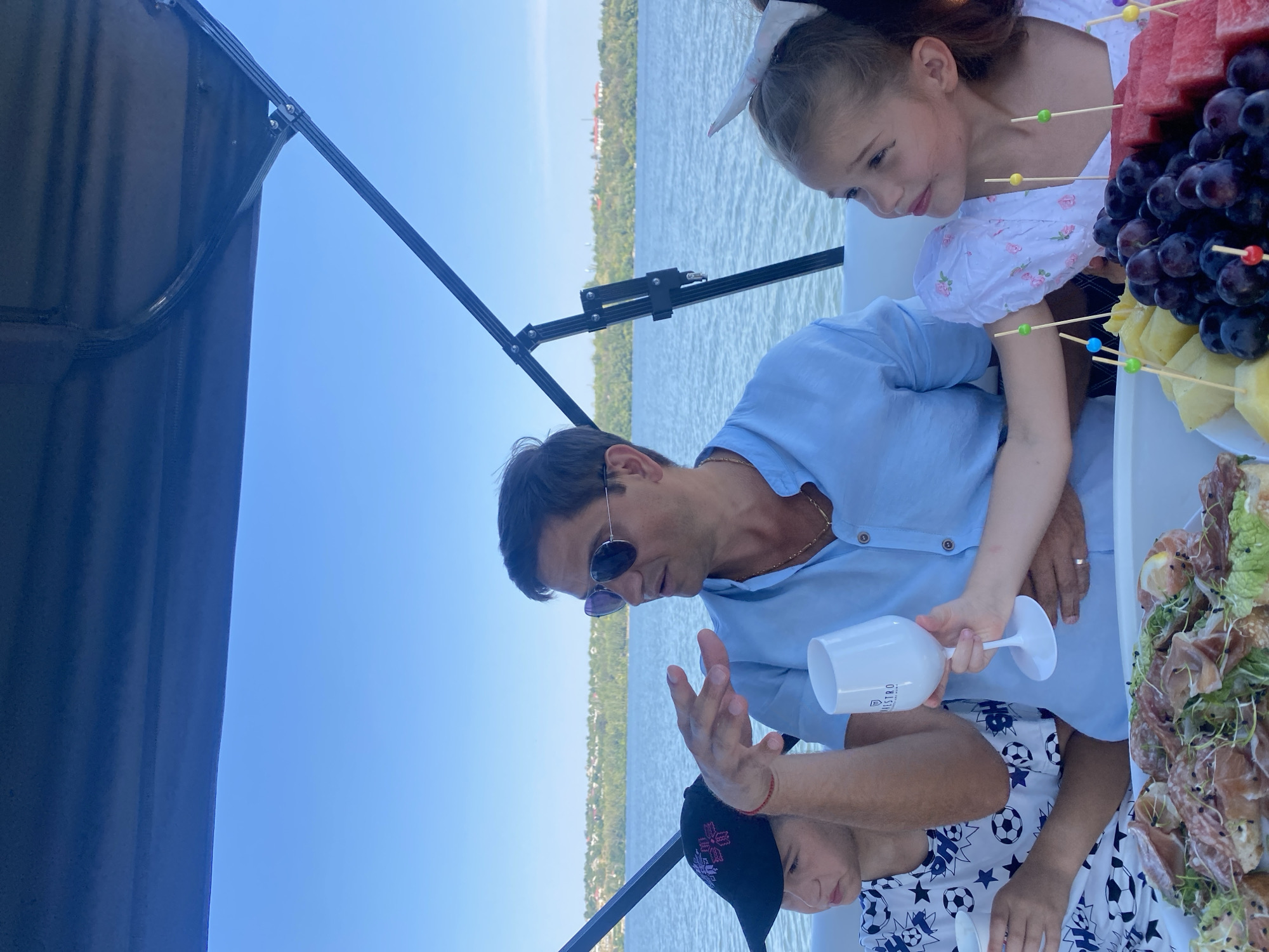 The image shows a group of people on a boat on a river. The water is calm and the sky is clear blue. There are trees and buildings on the distant shore. The people are dressed in casual summer clothing. In the foreground, there is a platter of food, including fruit and cured meats. A man in a light blue shirt is holding a wine glass and talking. A young boy in a black baseball cap is sitting next to him, and a young girl with a white bow in her hair is on the other side. The overall scene suggests a pleasant day out on the water.