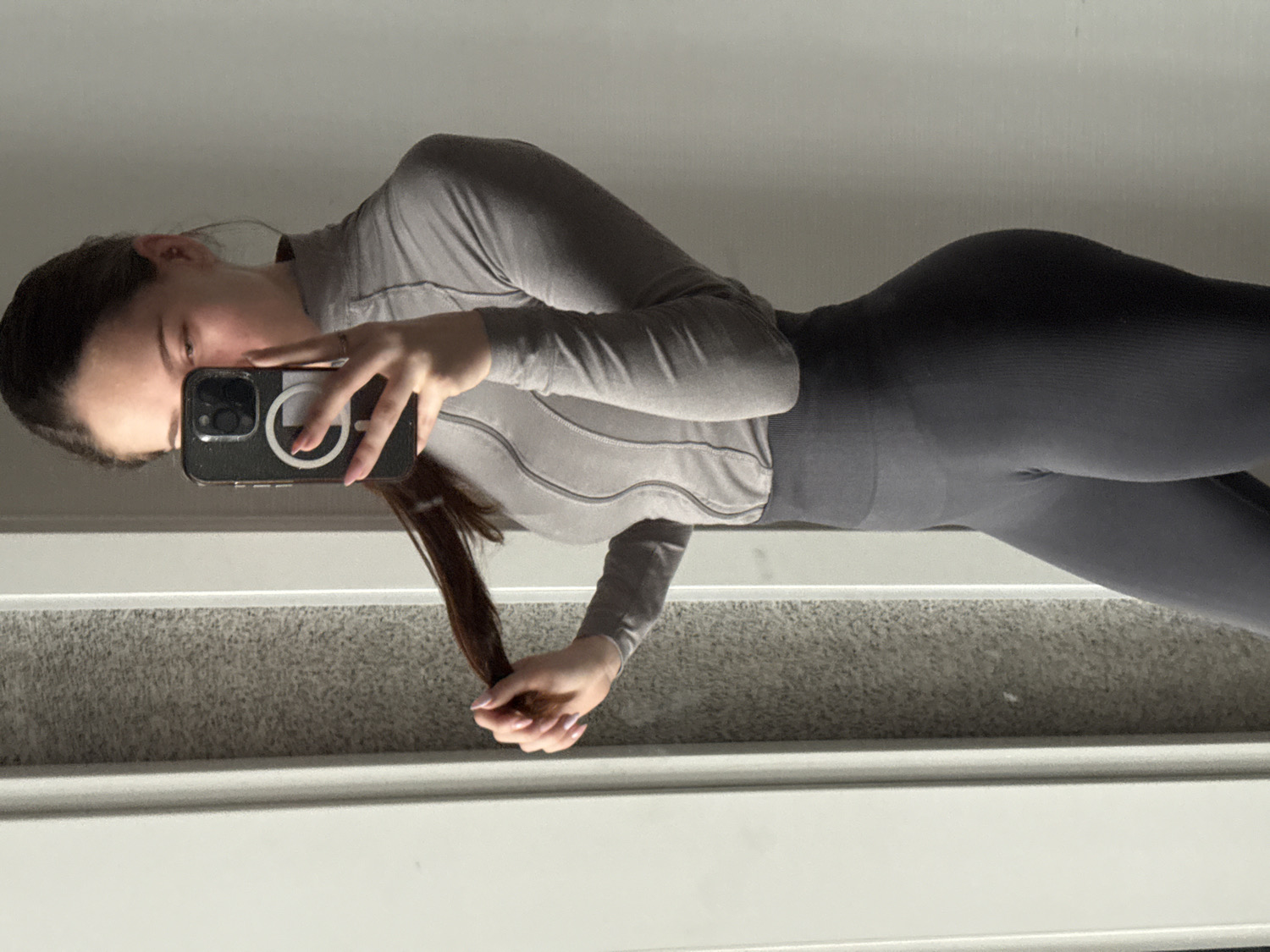 A young woman is captured in a mirror selfie, seen from the waist up. She has long, dark brown hair, mostly pulled back, with some strands framing her face. She is wearing a form-fitting athletic outfit consisting of a light grey long-sleeved top with subtle ribbed or textured details across the chest and arms, and dark grey high-waisted leggings. A thin ring is visible on her right hand, and her fingernails are painted with a light polish.
She holds a dark-colored smartphone in her right hand, which partially obscures her face as she takes the photo. The phone is in a black case with a white circular accessory, possibly a MagSafe attachment, prominently displayed on its back. This accessory features a white square outline with what appears to be a symbol or letter in its center. Her left hand is holding a section of her long hair.
The setting is indoors, appearing to be a room with a mirrored wall. The floor visible in the reflection is covered with light grey carpet. A white baseboard runs along the bottom edge of the wall or mirror. The background beyond the subject is light and indistinct. The lighting is soft and even, suggesting an indoor setting during the daytime. No specific location cues pointing to Sofia, Bulgaria, are visible in the image itself.