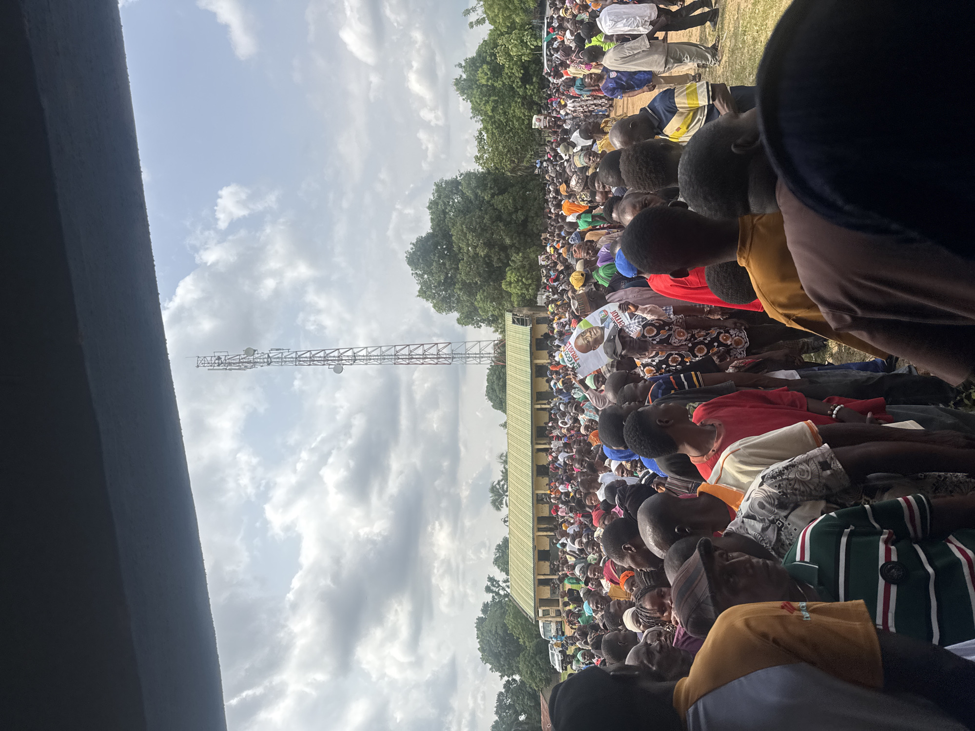 A large crowd of people are seated outdoors, likely in an open field in Akwanga, Nigeria. Many are wearing colorful clothing. A cell tower stands in the background. In the distance there is a building with a light-green roof and tall trees. The sky is cloudy.