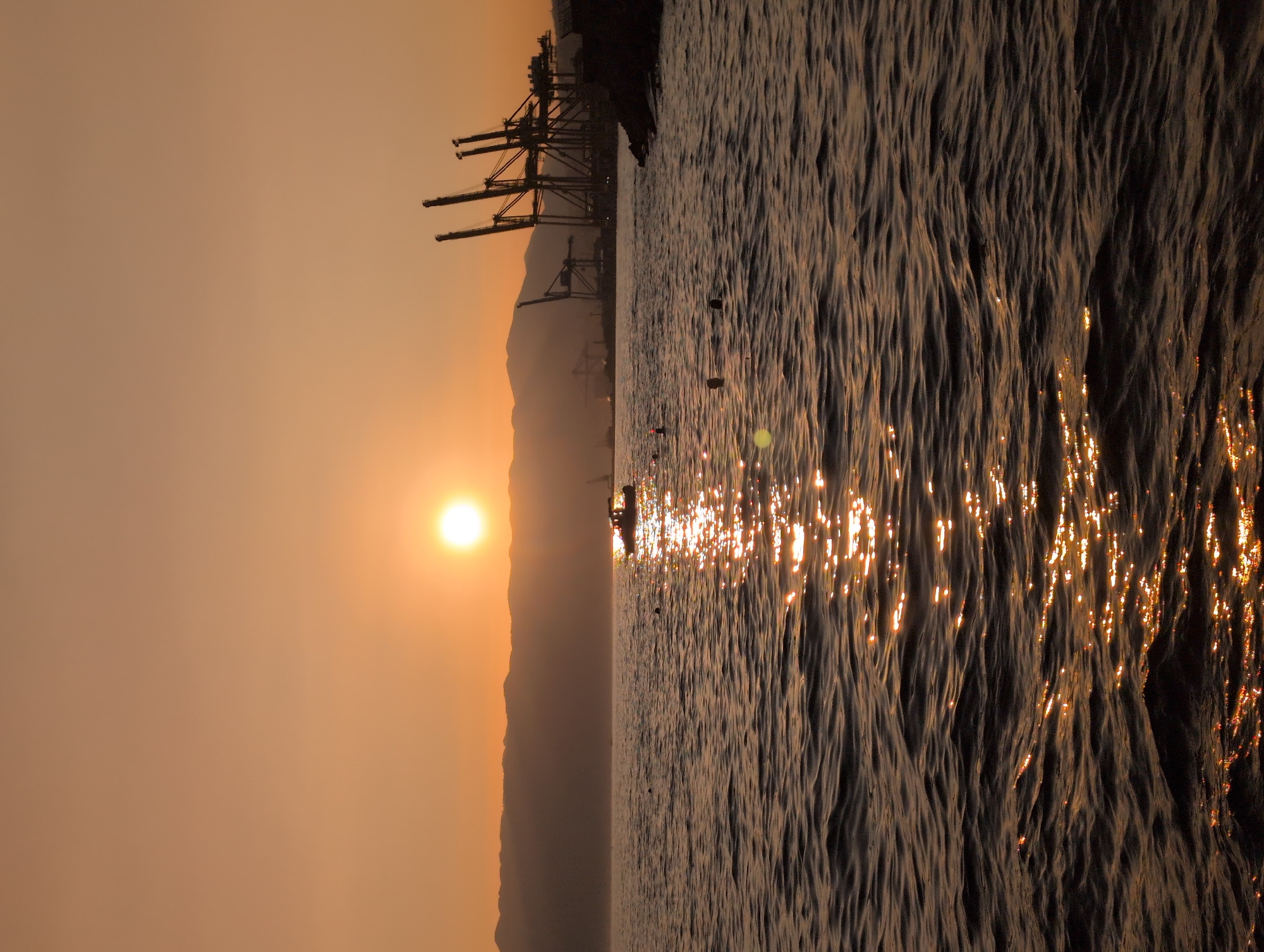 The image depicts a serene sunset over the water, possibly in the port area of Rijeka, Croatia, given the presence of industrial cranes visible on the horizon. The sun, a bright orb, is positioned in the upper left quadrant of the sky, casting a warm, orange glow across the scene and reflecting a shimmering path on the water's surface. The water itself is gently rippled, creating a textured foreground. In the middle ground, a small boat with occupants is visible, illuminated by the setting sun. Further in the distance, silhouetted against the sunset, are several large industrial cranes, characteristic of a port, suggesting an active harbor. Mountains form a hazy backdrop behind the industrial structures. The overall atmosphere is tranquil, with the warm light of the sunset dominating the color palette.