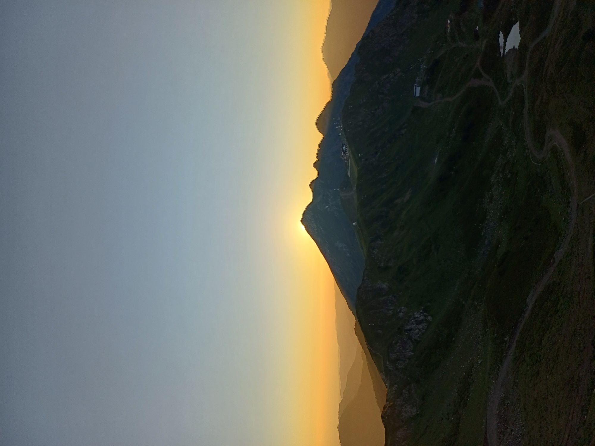The image captures a scenic mountain landscape at sunset. The sky is a gradient of soft blues and oranges, with the sun just peeking over the crest of a central mountain peak. Several layers of mountain ranges recede into the distance, fading into a hazy orange glow. The foreground features a steep, grassy mountainside with visible hiking trails and a small body of water. Structures, possibly resort buildings or observation points, are scattered on the slopes of the mountains. The scene appears to be a natural vista in the vicinity of Estosadok, Russia, known for its mountainous terrain and outdoor recreational activities. There are no people or animals visible in the image.