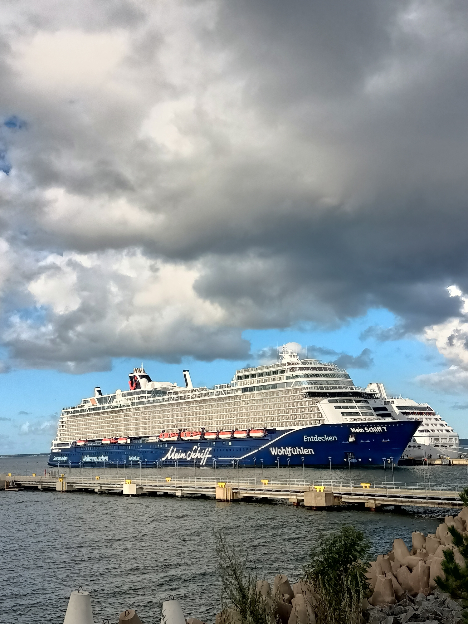 A large cruise ship, the Mein Schiff 7, is docked at a pier in Tallinn, Estonia. The ship is predominantly white with a dark blue hull that features the German text "Mein Schiff 7" and other German phrases like "Wohlfühlen" and "Entdecken". Several lifeboats are visible along the side of the ship. The sky above is filled with dramatic, dark grey and white clouds, with patches of blue sky visible. The pier extends out into the water, with lampposts lining its length and concrete barriers visible in the foreground along the shoreline. Another cruise ship is partially visible behind the Mein Schiff 7. The water is choppy. There are no people visible on the ship or the pier.