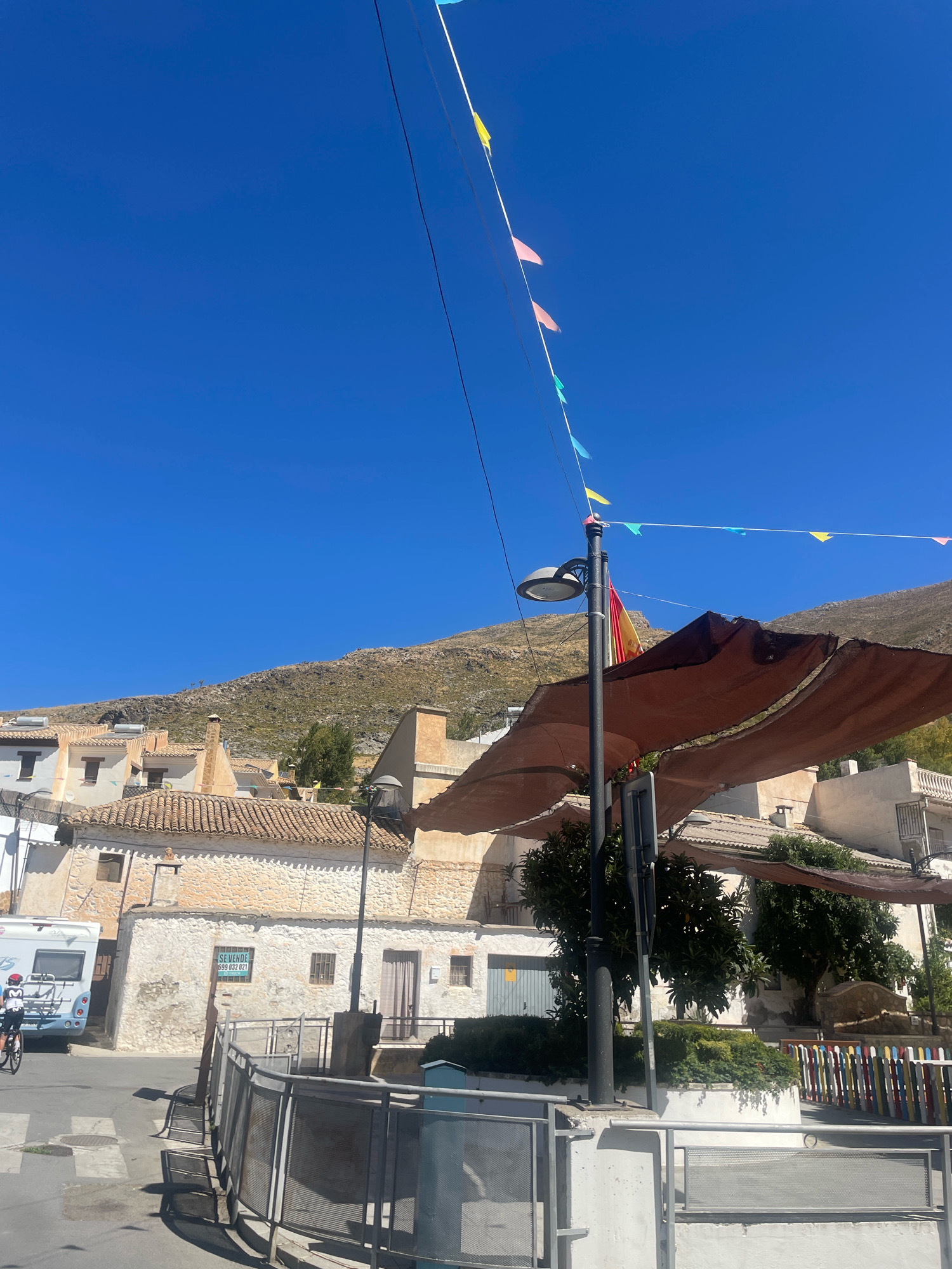 The image depicts a street scene in Güéjar Sierra, Spain, under a clear blue sky. In the foreground, a metal fence with a gray railing encloses an area with greenery. A lamppost with a circular lamp head stands to the right, adorned with a Spanish flag. A brown awning stretches overhead. Behind the fence, white buildings with tiled roofs are visible, some appearing aged with textured walls. To the far left, a white RV is parked. A person on a bicycle, wearing a red helmet and white shirt, is riding past the RV. A sign on one of the buildings reads "SE VENDE" (For Sale) with a phone number. In the background, a rocky, barren hillside rises, with more buildings dotting its lower slopes. Colorful bunting flags are strung across the sky, adding a festive touch to the scene. The overall impression is of a sunny day in a Mediterranean town.