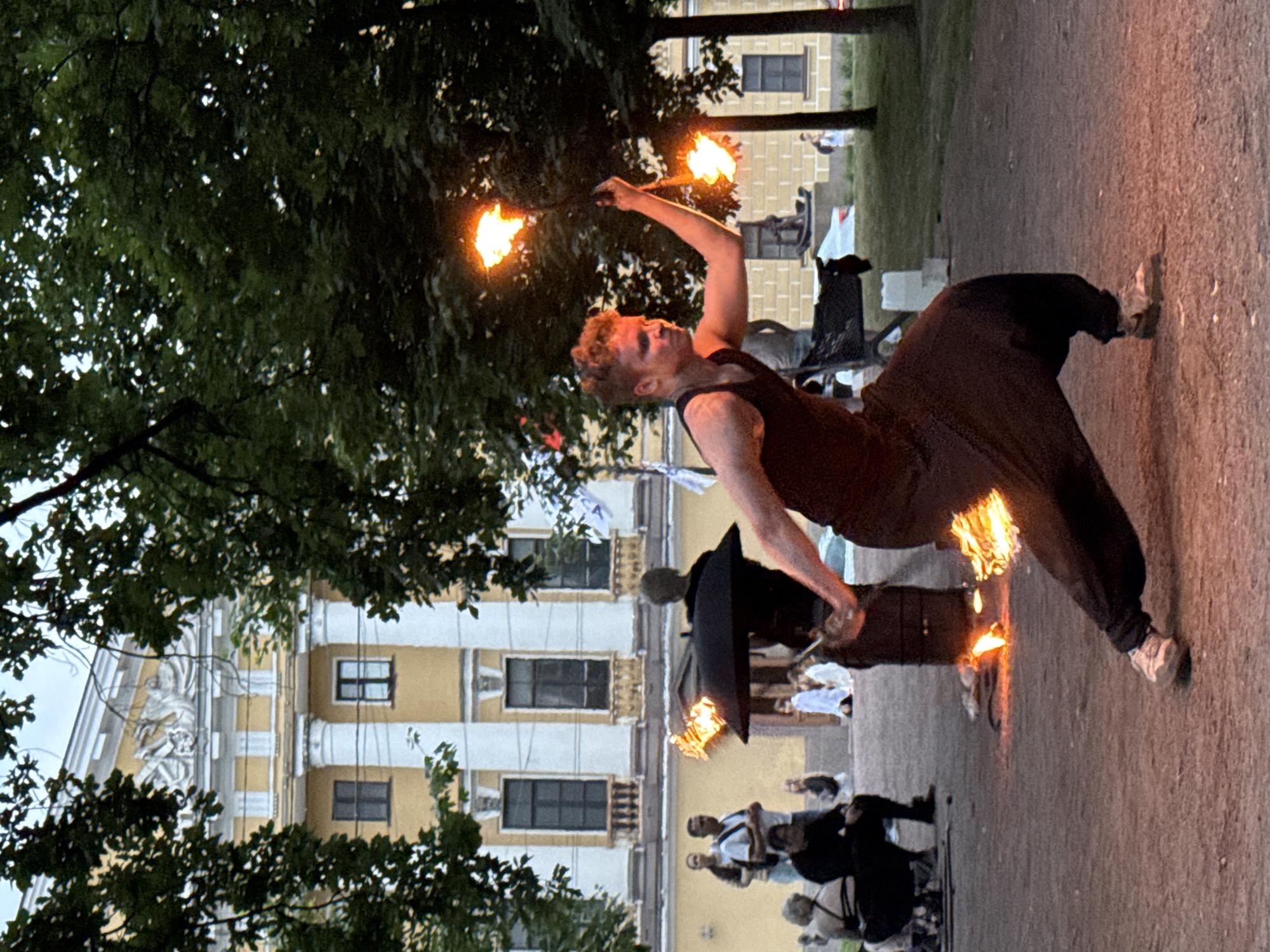 This image captures a street performer engaged in a fire-dancing demonstration on a warm evening. The performer, a young male with reddish-blond hair, is wearing a dark tank top and loose-fitting dark pants. He is actively twirling fire props, with flames emanating from them, creating a spectacle of light and movement.
In the background, a grand, pale yellow building with white columns and ornate architectural details stands prominently. This building appears to be a significant historical or cultural landmark, likely a museum or a government institution. The surrounding environment suggests a public square or park area within the city of Saint Petersburg, Russia, given the classical architecture typical of the city.
Several onlookers are present, observing the performance from a distance. They are scattered around the square, some seated, others standing. The overall atmosphere is one of casual entertainment and public engagement. The lighting suggests the late afternoon or early evening, with a mix of natural light and the illumination from the fire props. The ground is a dusty, earthen surface, common in outdoor public spaces. Trees with dense green foliage frame the scene, adding to the ambiance of an outdoor performance in a pleasant urban setting.