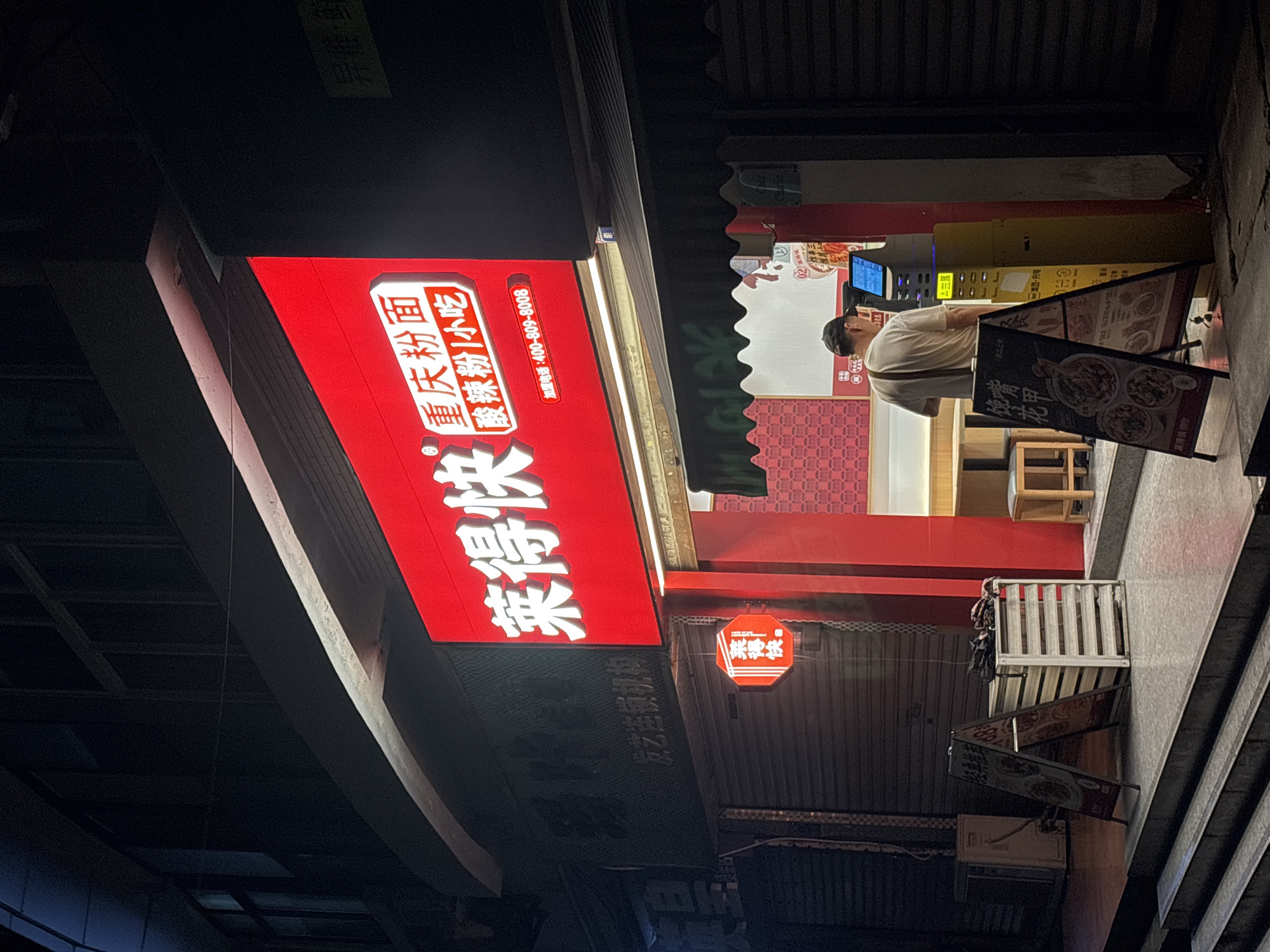 The image shows a nighttime view of a street scene in Nan'an District, China. The focus is on the storefront of a restaurant called "莱得快" (Lái dé kuài), which is also branded as "重庆粉面" (Chongqing Noodles) known for "酸辣粉小吃" (Suan La Fen Small Eats). The storefront is identifiable by its red signage with white Chinese characters. A man is ordering at a self-ordering kiosk. Menu boards are visible.