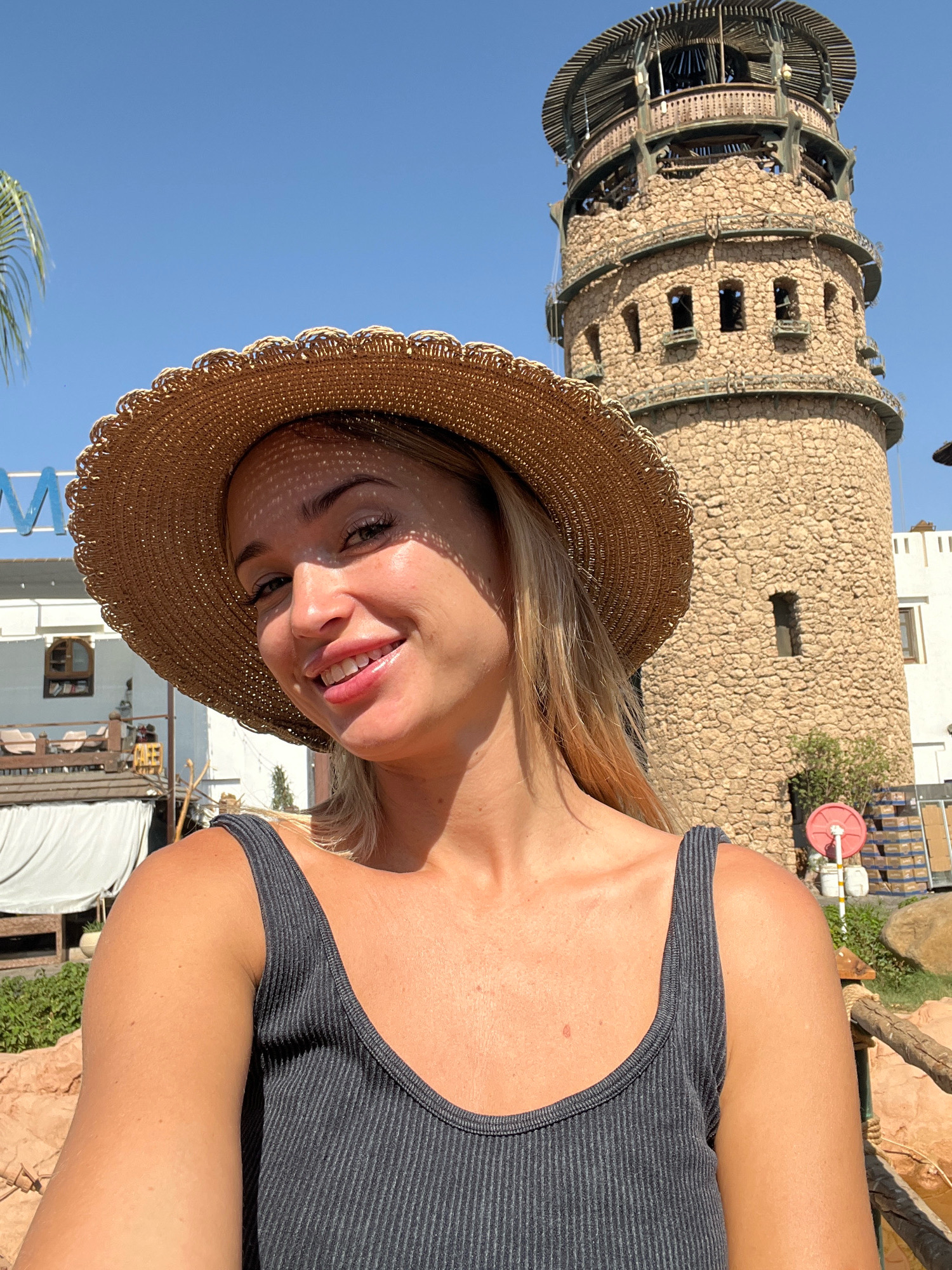 A young Caucasian woman is taking a selfie outdoors with a straw hat on and a gray tank top. She is smiling. Behind her, a tall, round stone tower with multiple levels and open windows stands against a clear blue sky. There are some buildings and vegetation in the background, suggesting a resort or tourist area. The lighting indicates it is daytime. The overall scene appears to be in Sharm El Sheikh, Egypt, possibly at a resort or attraction like a lighthouse or observation tower.