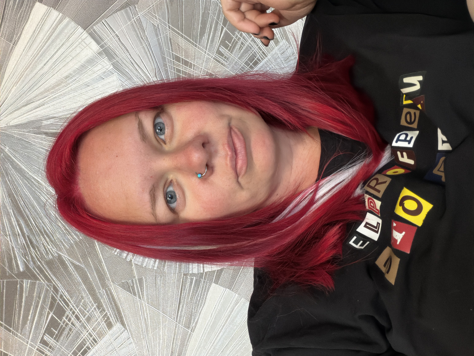 The image is a vertical, close-up selfie of a person with vibrant red hair. The individual has light blue eyes and a nose ring with a small blue gem. They are looking at the camera with a slight smile. The person is wearing a black t-shirt with colorful block letters that appear to spell out "EL PROFESOR". The background is a textured wallpaper with a repeating pattern of radiating lines and shapes in shades of gray and white. The lighting suggests it is daytime, and the indoor setting is illuminated by artificial light. The subject's hand is visible at the top of the frame, with black nail polish. The overall mood is casual and relaxed.