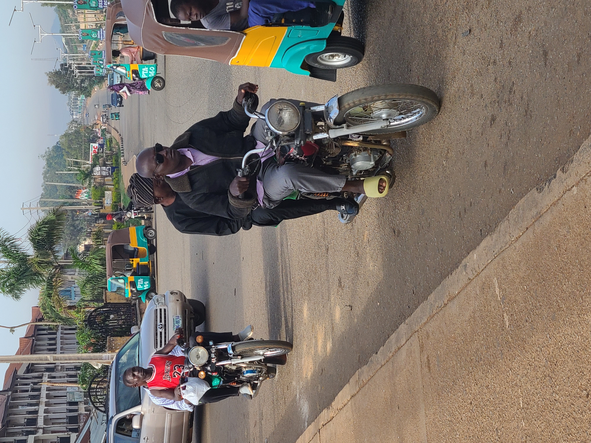 The image depicts a street scene in Gura Top, Nigeria. In the foreground, a man wearing sunglasses and a black jacket sits on a vintage motorcycle. He appears to be the driver. A second person sits behind him, partially obscured. The man on the motorcycle has one foot on the ground, while the other rests on the motorcycle. The street is paved and dusty. A yellow and teal "keke" or auto-rickshaw is visible in the upper right corner of the image, and others are farther away in the opposite direction. A silver-colored SUV is parked on the left side of the image. Another man is on another motorcycle, parked in front of the SUV. The background shows a mix of buildings and vegetation, with billboards lining the street. The sky is hazy.
