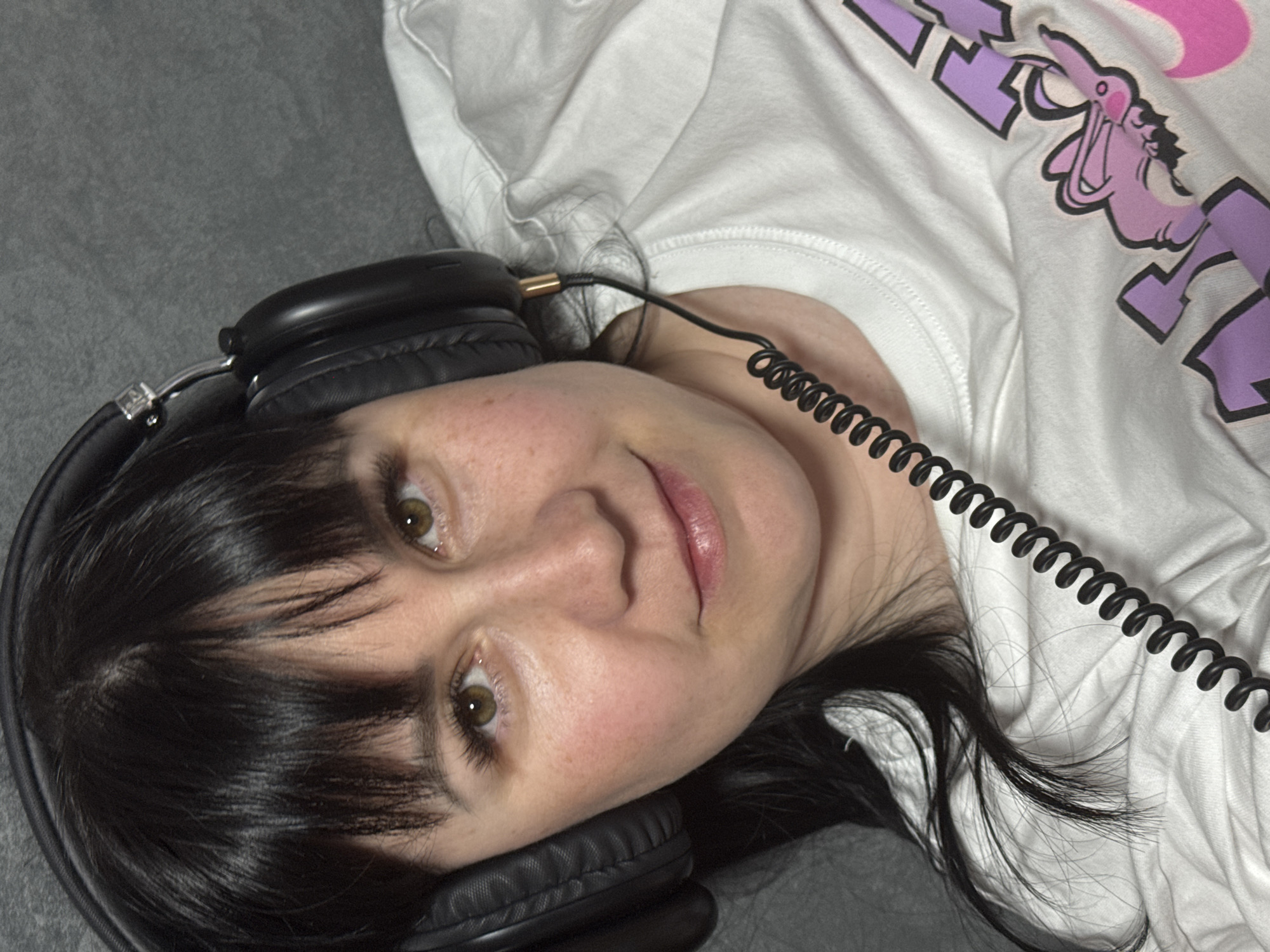 A person is lying down with their head tilted to the left, wearing black headphones with a coiled black cord. They have dark hair and are wearing a white t-shirt with a pink and purple design. The background is a gray surface. The location in Zav'yalovo, Russia is unknown.