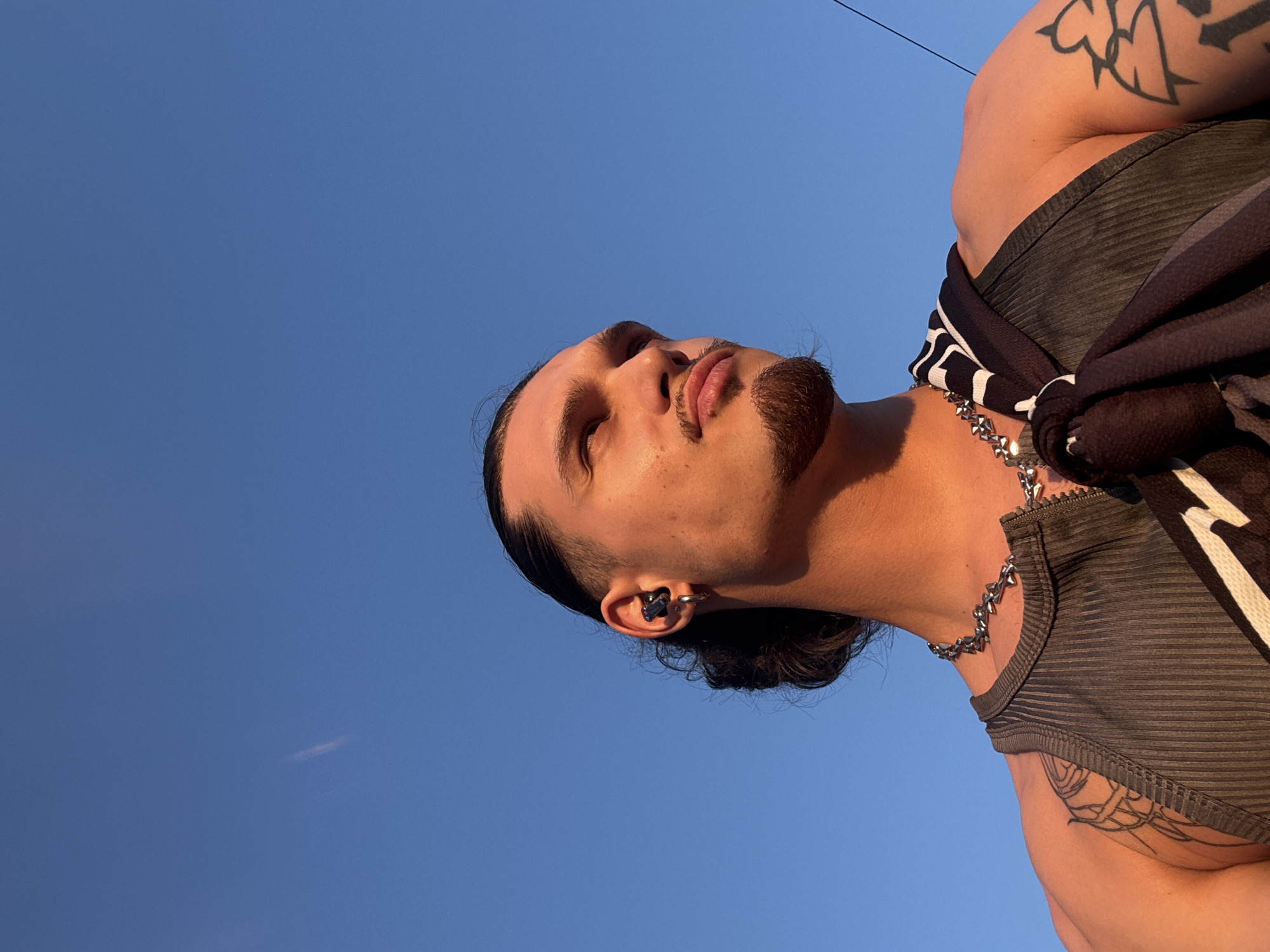A low-angle, close-up outdoor shot features a young man looking up and to his right against a clear blue sky. He has dark hair pulled back into a bun, a goatee, and a mustache. He is wearing a grey ribbed tank top, a chunky silver chain necklace, and a dark bandana with white stripes draped over his left shoulder. An earbud is visible in his right ear, and a hoop earring is in his left. Tattoos are visible on his left shoulder and arm. A faint white streak, possibly a contrail, is visible in the sky. The lighting suggests it is either early morning or late afternoon. No specific location in Sofia, Bulgaria can be identified from the image.