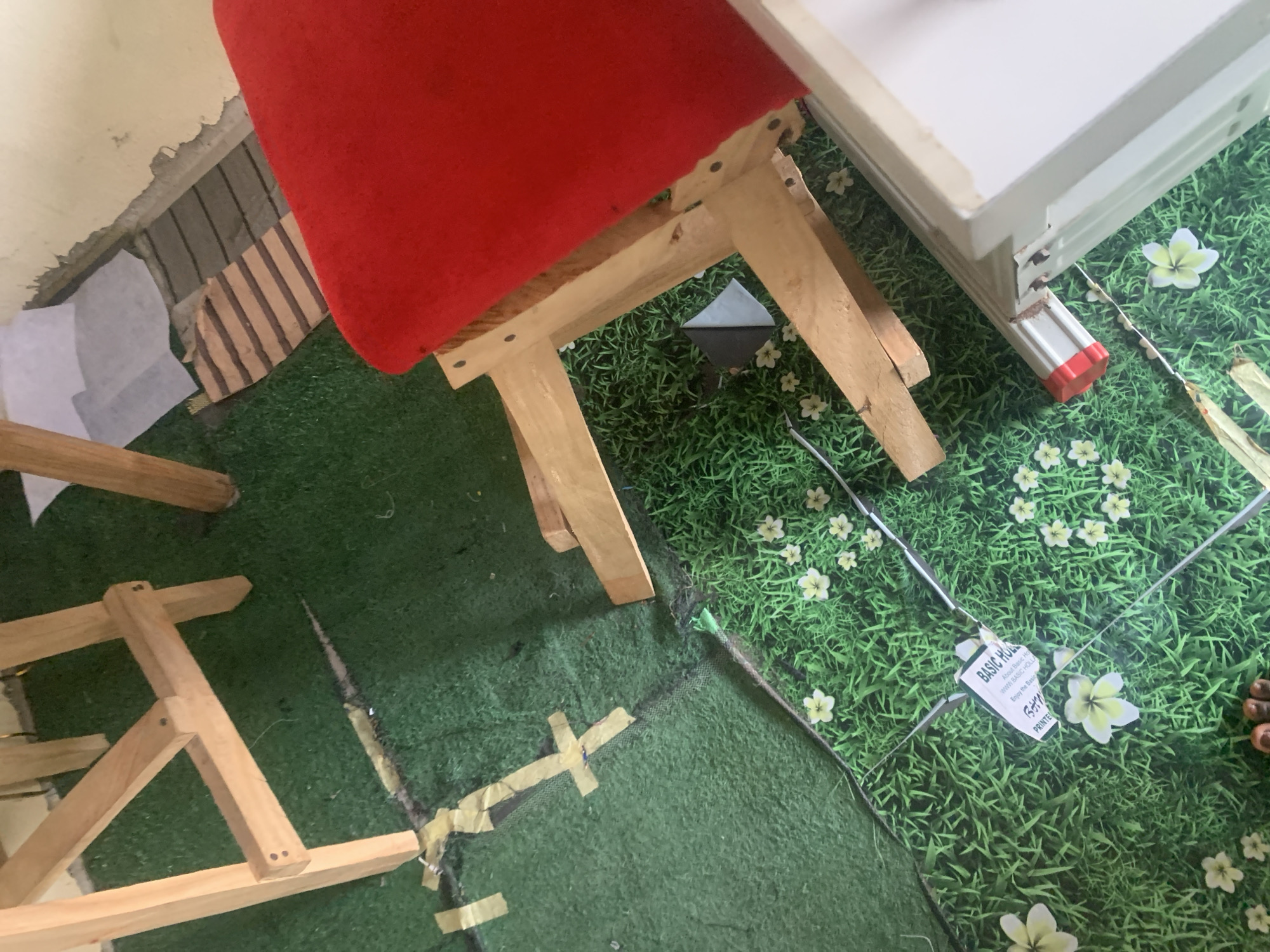 This image depicts an indoor scene, possibly a room, located in Jauro Yinu, Nigeria. The floor is covered with artificial grass and a printed grass mat with flowers. Two wooden structures, one appearing to be part of a table or chair frame with a red seat, and another simpler wooden structure, are present. A small black pyramid-shaped object is visible on the floor. A label with text, including "BASIC HOL..." and "PRINTED...", is also on the floor. The scene has a somewhat unfinished or temporary appearance, with visible tape on the artificial grass and some exposed construction materials along the wall. No individuals are clearly visible, though a portion of what appears to be feet in the lower foreground suggests human presence.