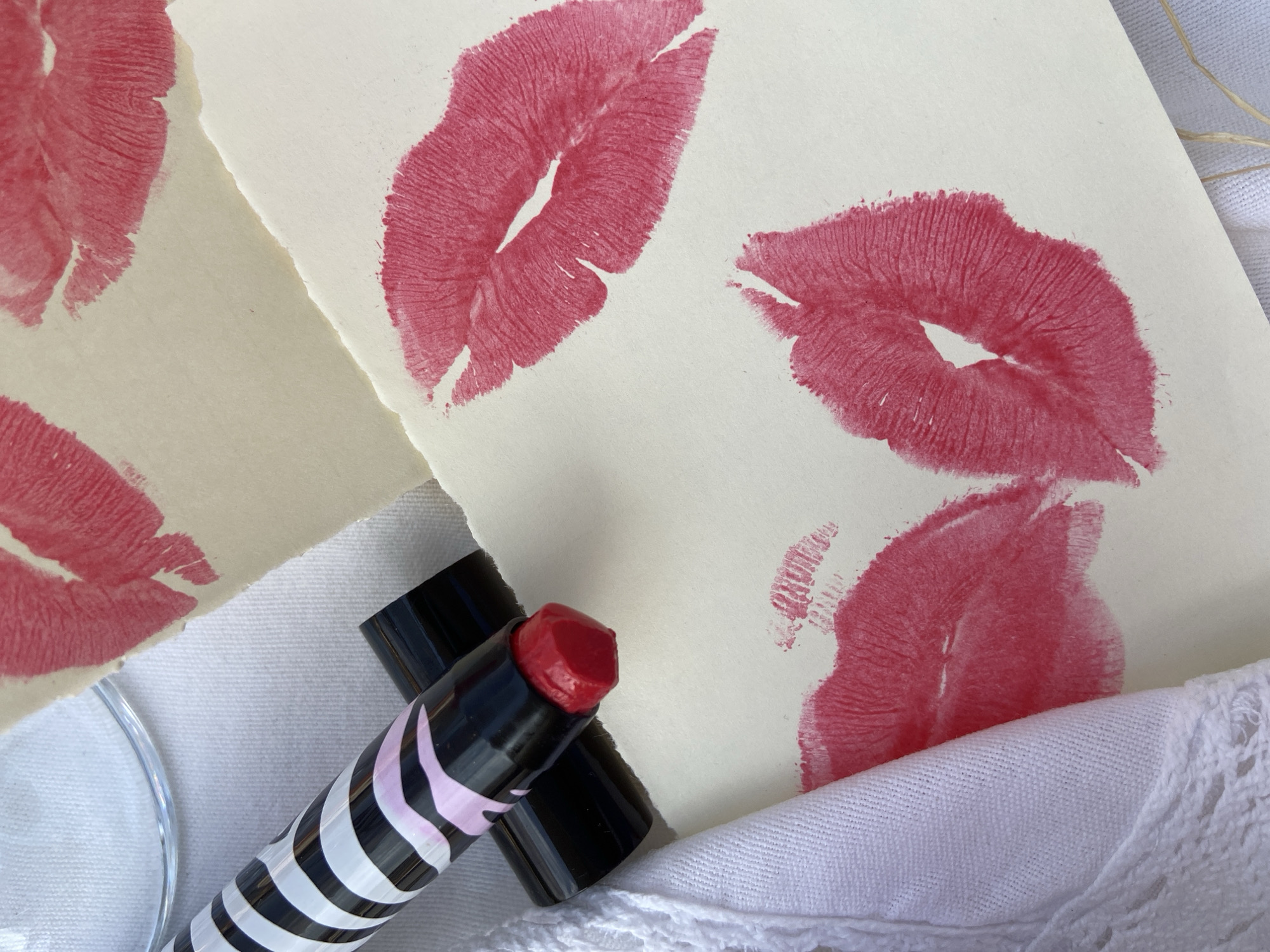 The image shows a close-up still life arrangement featuring two pieces of paper with red lipstick kiss prints, a tube of red lipstick, and white fabric.
The paper pieces have torn edges, suggesting they were intentionally ripped. Several red lipstick kiss marks are visible on both pieces of paper. A tube of lipstick with black and white stripes and a pink stripe near the top is situated near the paper. The cap of the lipstick is off, revealing the red lipstick. The arrangement is set on a white fabric with a lace trim. The environment appears to be a flat surface, possibly a table, photographed in natural light. The location of Budva, Montenegro, is not evident in the image and cannot be determined from the visual information provided.