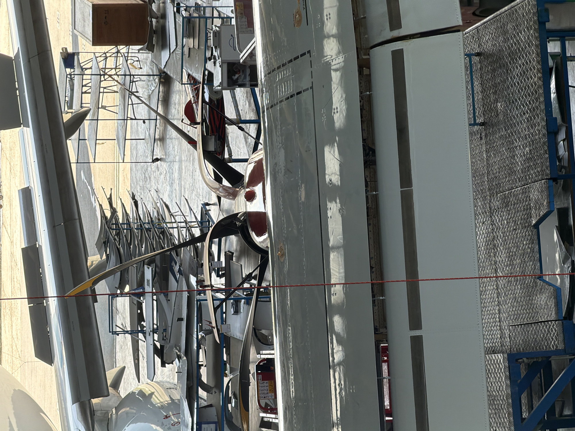This media file captures an industrial interior, specifically an aircraft maintenance or manufacturing facility, likely a hangar, located in Sofia, Bulgaria. No people are visible in the image.
The scene is dominated by large sections of aircraft, primarily fuselages or wings, suggesting ongoing assembly, maintenance, or storage. In the foreground and center-right, a long, light grey aircraft section with visible riveted panels and some dark grey markings extends across the frame. Another large, white aircraft component, possibly a nose cone, is partially visible in the bottom-left corner.
Numerous smaller, elongated aircraft components, resembling propeller blades or control surfaces, are arranged vertically on metal racks in the mid-ground and background, indicating a well-organized storage or staging area. Various metal scaffolding, blue-painted support structures, and industrial equipment are scattered throughout the facility. A blue metal cart with a diamond-plate surface is visible on the far right. A red string or cable traverses horizontally across the middle of the image.
Other objects include a large wooden crate in the upper background with some illegible text, and a red piece of safety equipment, possibly a fire extinguisher, near one of the aircraft sections. The lighting suggests an indoor environment, with reflections on the shiny aircraft surfaces indicating overhead artificial lights and possibly some natural light.
Visible text is limited and mostly illegible due to resolution or angle. Faint markings like "NO STEP" are discernible on one of the dark grey aircraft components in the left background, and some indistinct characters appear on the wooden crate. The overall atmosphere is functional and industrial.
