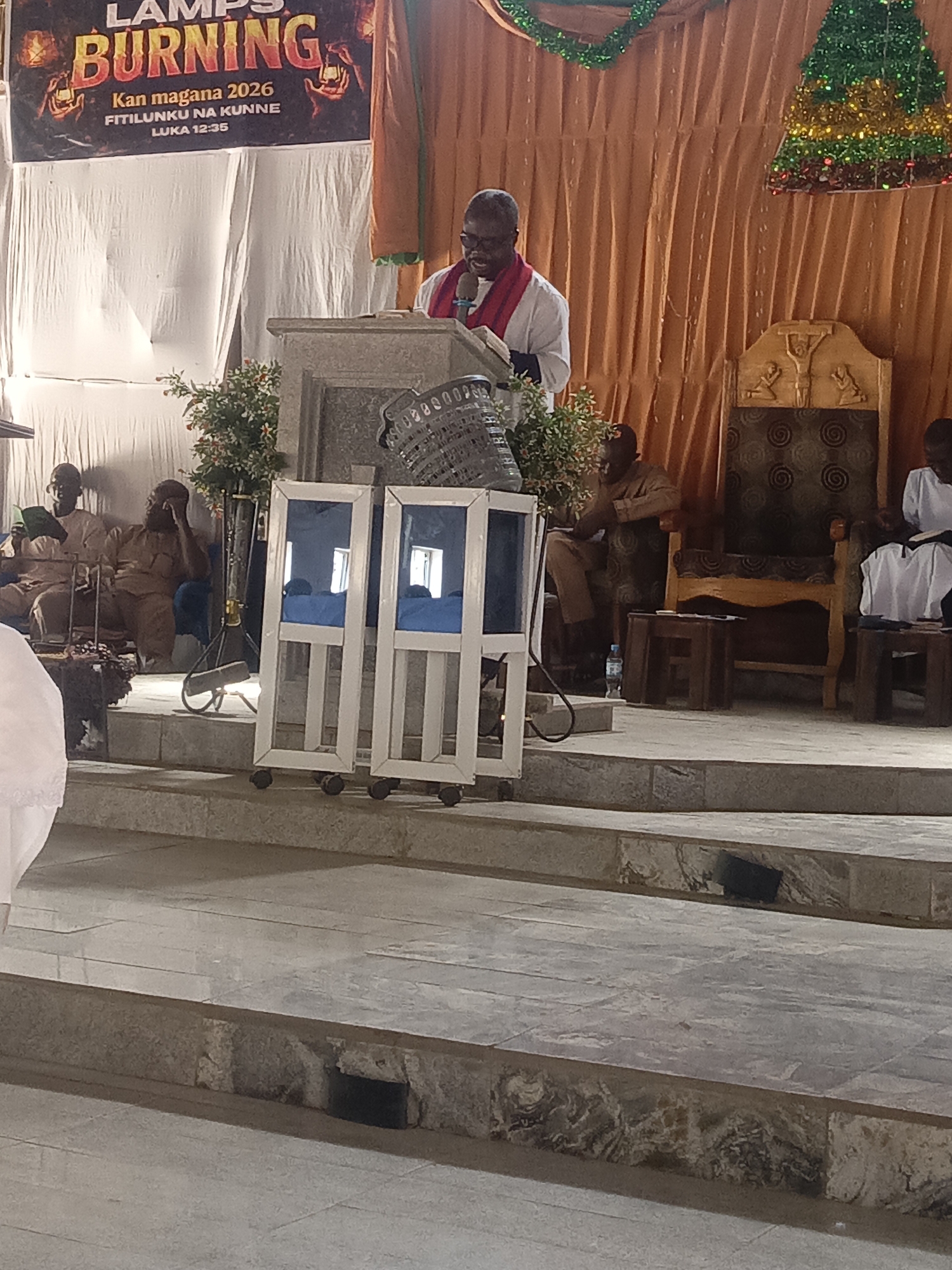 A church in Unknown, Unknown features a speaker at a podium with a "LAMPS BURNING" banner overhead. The interior includes a gold-curtained backdrop and a decorative Christmas tree. Congregation members are seated on a raised platform with chairs and stools, alongside portable partitions on wheels. The room has stone-tiled steps leading to the platform.