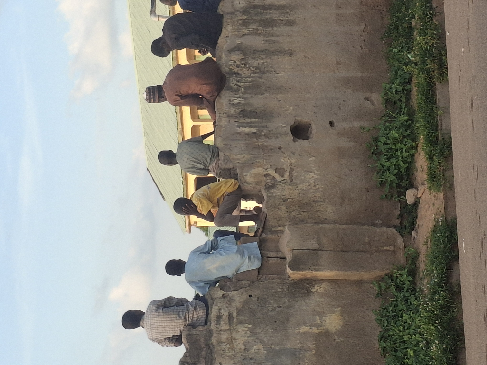 A group of six young men are sitting on a low, weathered stone wall in Pantame, Nigeria. The wall appears to be part of an old structure, possibly a historical building or a section of the city wall. The men are positioned from left to right, facing away from the camera. The man on the far left wears a checkered shirt. Next to him, a man in a light blue garment sits with his back to the viewer. The third man from the left is in a yellow shirt, with his hand to his face. The fourth man, wearing a brown garment and a cap, is also seated. The fifth man from the left is in a dark brown garment, and the sixth man on the far right is largely obscured, wearing a dark garment. Behind the wall, a building with a green roof is visible. The ground in front of the wall is covered with green vegetation, and a paved road is in the foreground. The sky is a light blue with scattered clouds, indicating it is likely daytime. The overall scene suggests a moment of rest or observation in an urban setting.