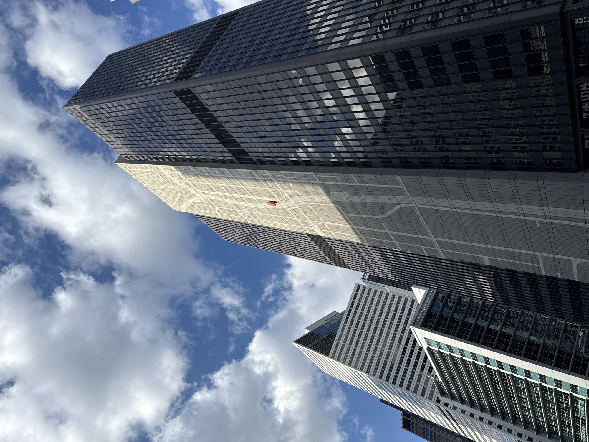 The photograph is taken from a low angle, pointing upward towards a cluster of tall buildings in Chicago, Illinois, United States of America. The blue sky, dotted with fluffy white clouds, serves as a backdrop. The primary building is dark, possibly black or dark gray, with a grid-like pattern suggesting windows. A portion of the building reflects the cloudy sky, creating a mirror-like effect. To the side of the dark building is a lighter-colored structure, featuring a detailed grid pattern. Near the bottom of the frame, parts of other buildings with similar grid-like architectural designs are visible. The overall scene depicts a typical urban environment, specifically showcasing the impressive architecture of downtown Chicago.
