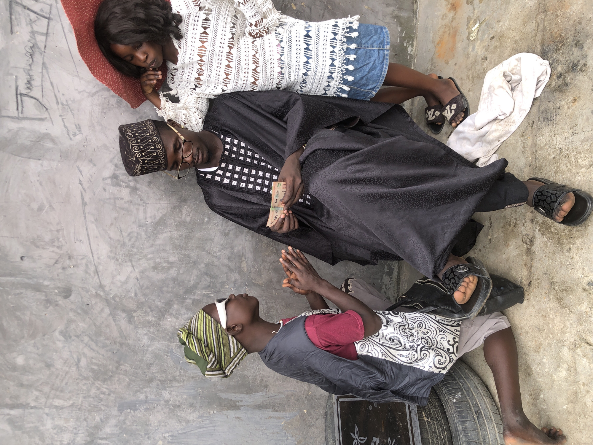 In Ado, Nigeria, a young person in a green and white striped headwrap and white sunglasses kneels on the ground, hands clasped. They wear a maroon t-shirt, a dark grey vest, and light-colored pants, with a patterned bag at their side. To their right, a man in a black cap and glasses, dressed in a black, embroidered robe, sits with money in his hands. Partially behind him, a young person wearing a white, lacy top and a denim skirt leans against a red object, looking downwards. The scene takes place against a concrete wall, with discarded tires and a crumpled white cloth on the ground.