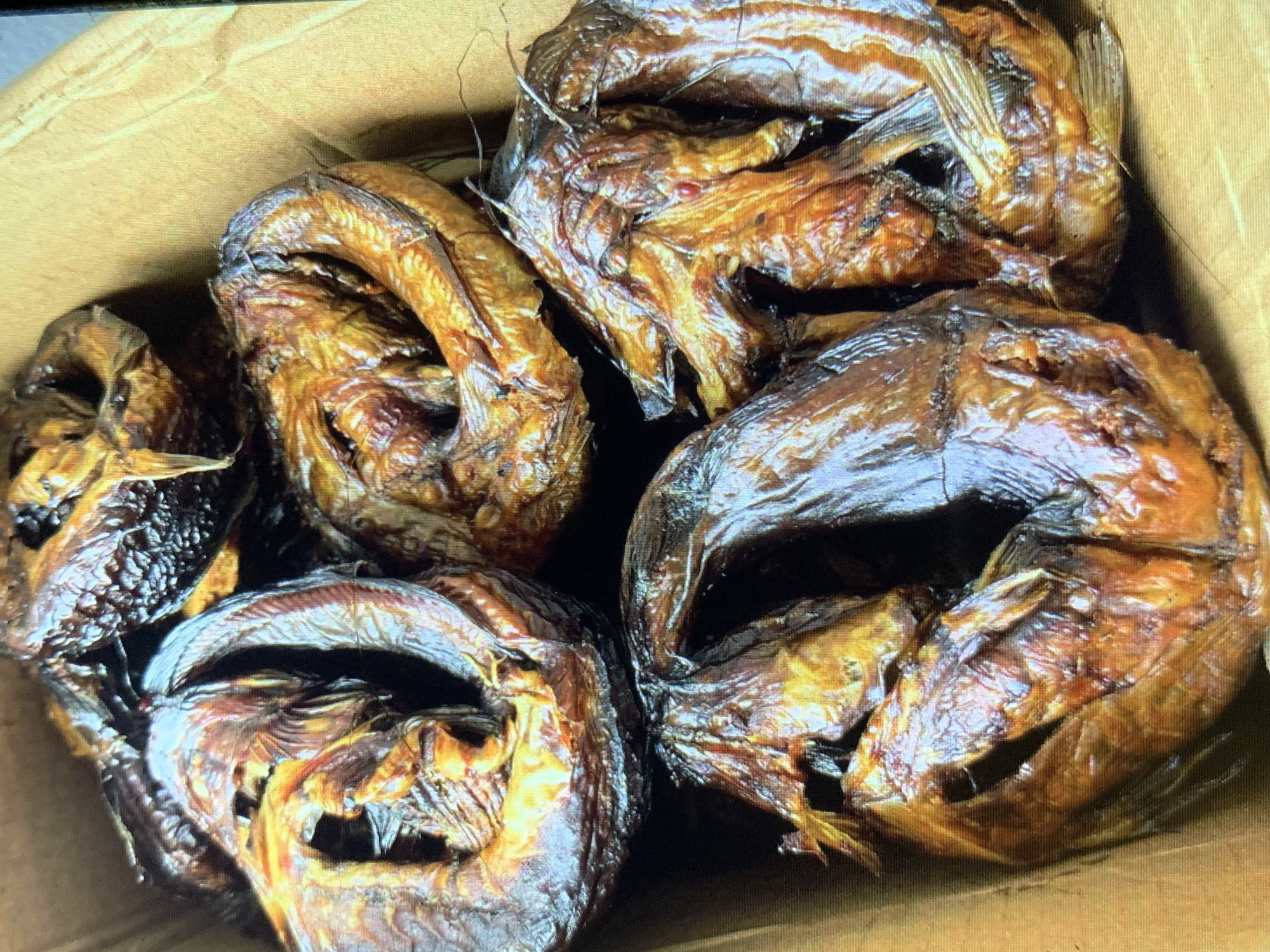 The image shows a close-up of smoked fish in a cardboard box. There are approximately six whole fish, arranged in a heap. The fish are dark brown, with visible scales and fins. The heads of the fish are curled, with open mouths. The box appears to be plain and unbranded.
It is not possible to determine if this image was taken in the city of Rafin Par, Nigeria.