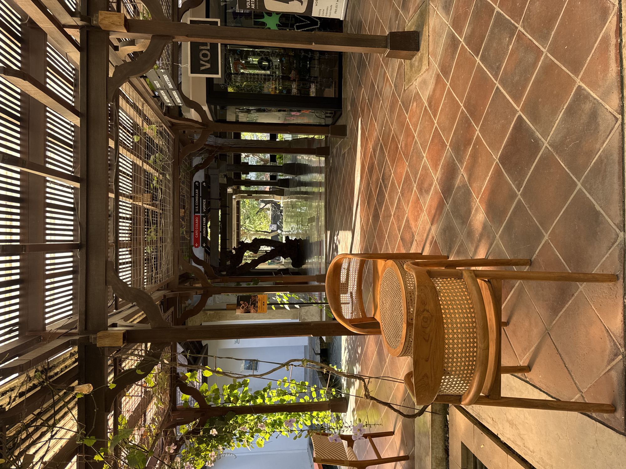 This image depicts an outdoor seating area with a thatched roof and wooden beams, located in Kuta Selatan, Indonesia. In the foreground, two light-wood chairs with woven seats and backs are positioned on a terracotta tile floor. A small round table sits between them. Lush green vines with small purple flowers hang from the overhead structure, casting dappled shadows. To the left, a narrow water feature is visible.
In the background, a walkway extends into the distance, lined with more wooden pillars and overhead structures. Signs indicate directions to "Cafe Colabo," "U.S. Consular," "Jungle Park," and "Voffice." Further down the walkway, a shopfront with the sign "VOL" is visible, displaying various items on shelves. The walkway leads to an open area with a body of water and what appears to be a large, dark sculptural tree. The scene is bathed in warm sunlight, suggesting it is daytime. No people are present in the frame.